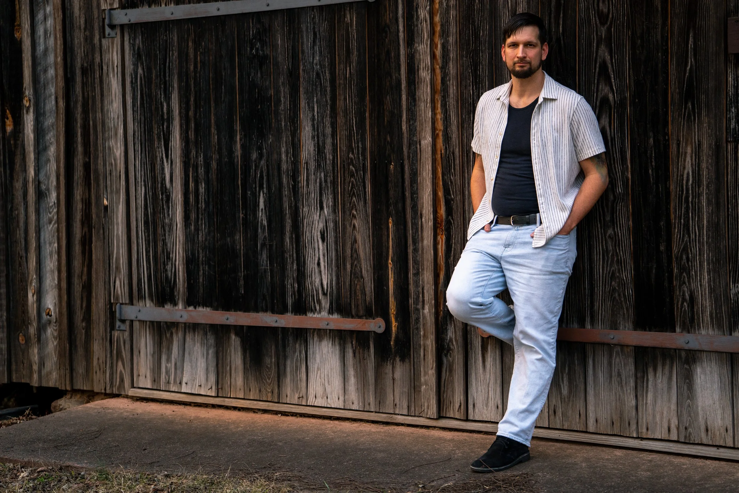 A young man in a white and black top and blue jeans poses in front of a rustic wood mill at West Point on the Eno Park, Durham North Carolina for High School Senior Photos
