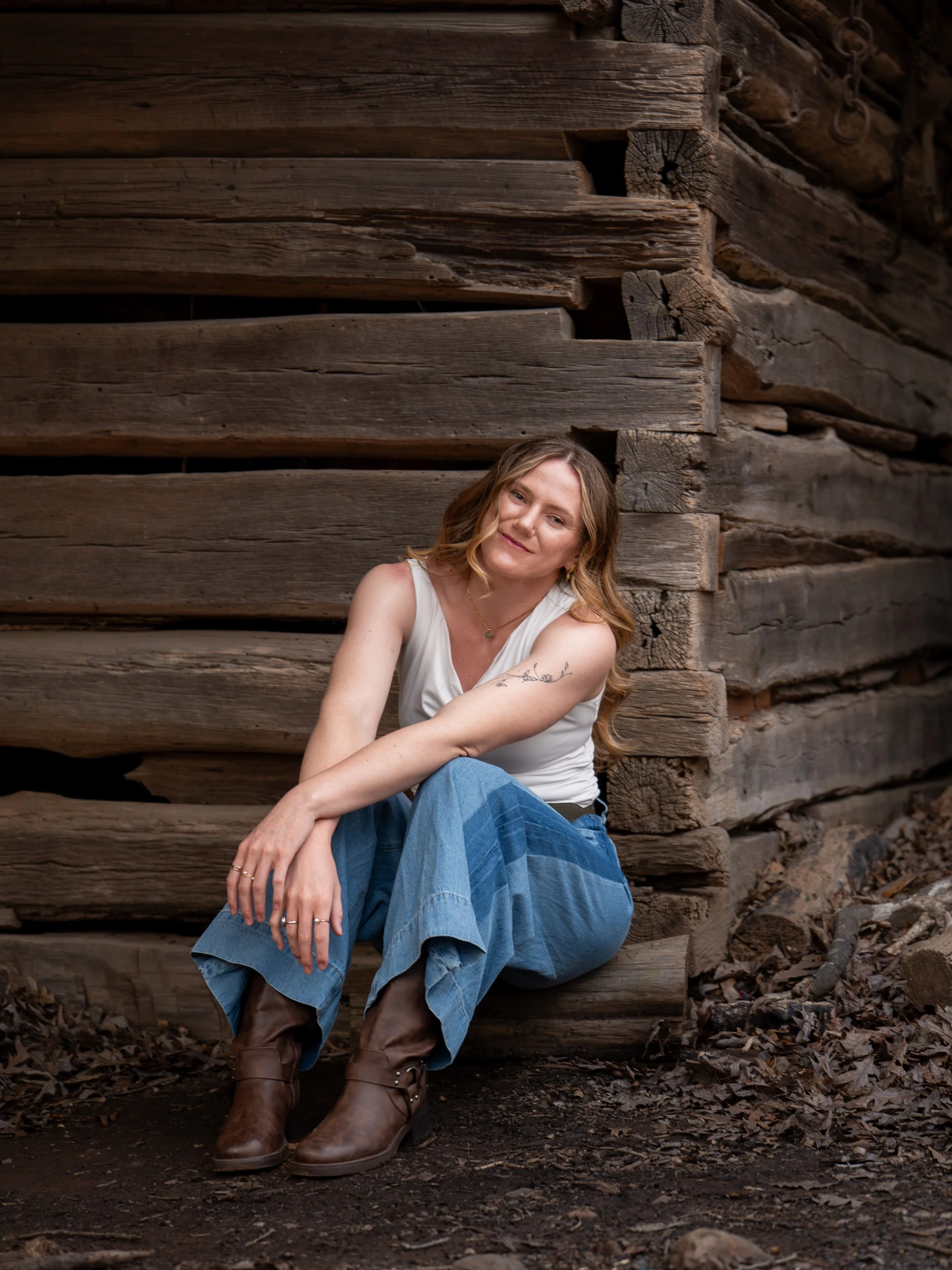 A young woman in a white top and blue jeans poses in front of a rustic wood mill at West Point on the Eno Park, Durham North Carolina for High School Senior Photos