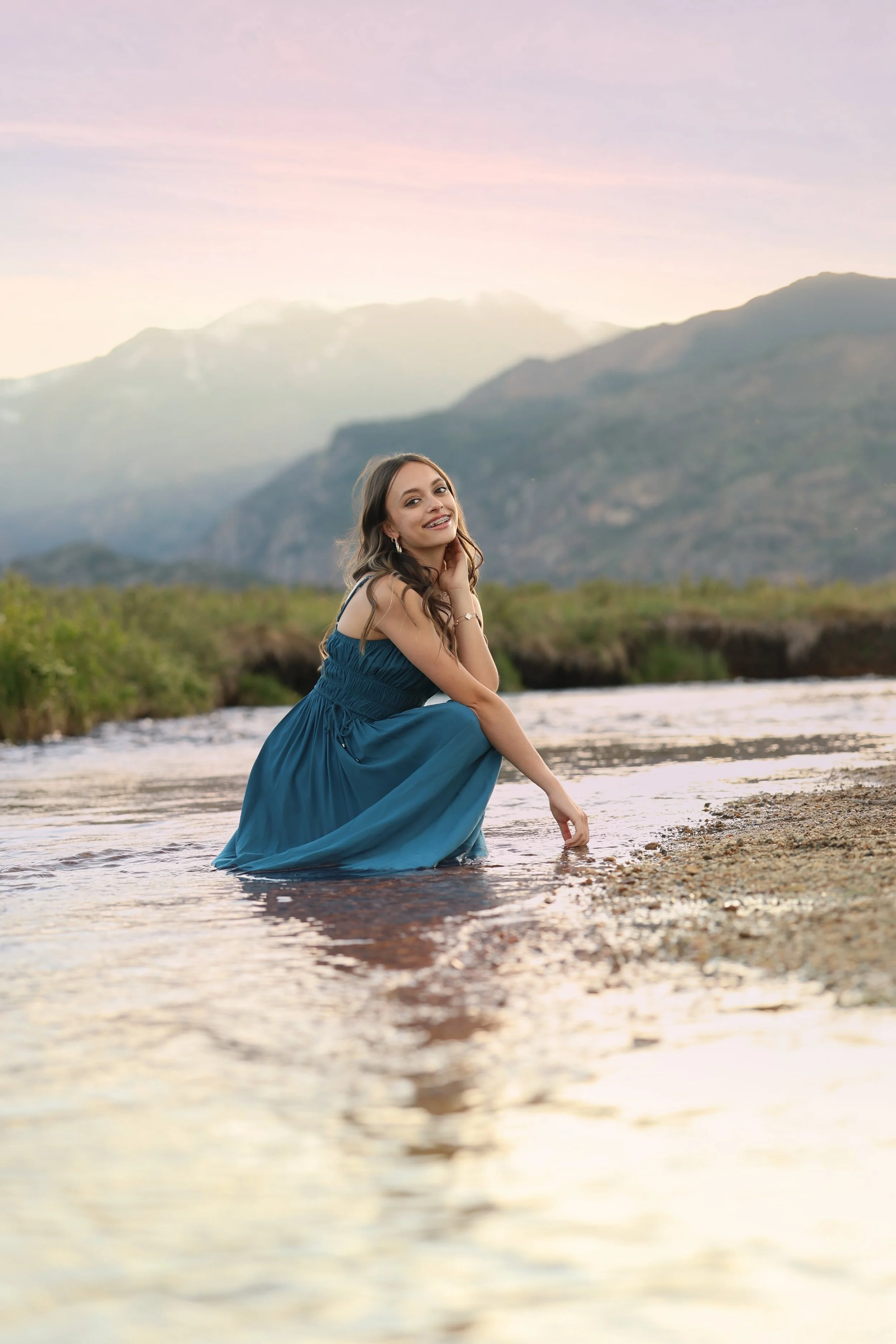 A woman wearing a blue dress sitting in shallow water near a river or lake, with mountains and a pastel-colored sky in the background.