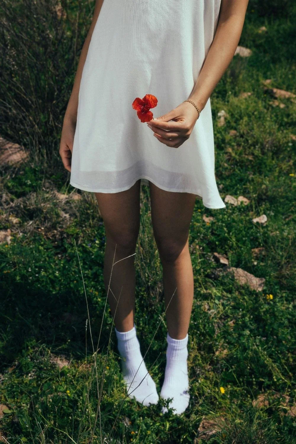 A woman wearing a white dress and white socks is standing on grass and holding a red flower.
