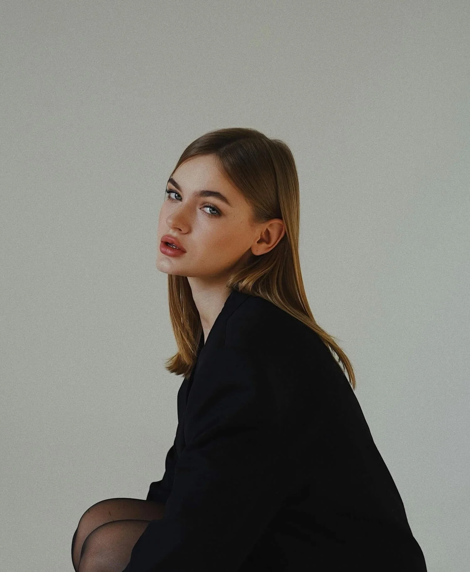 A woman with light brown hair and blue eyes sitting sideways against a plain gray wall, wearing a black blazer and sheer black stockings.