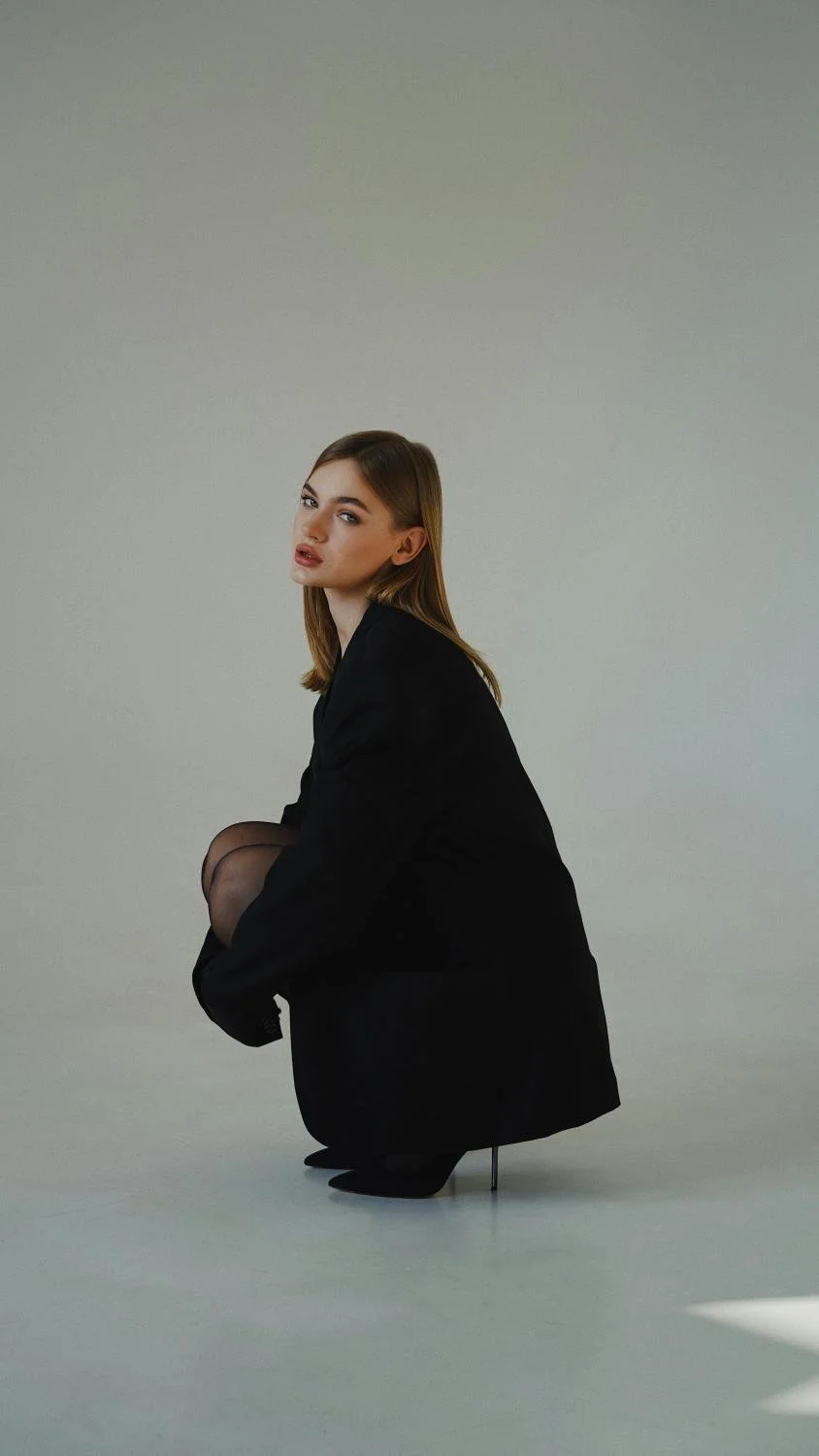 A young woman with brown hair, dressed in a black blazer and stockings, is squatting against a plain light-colored background.