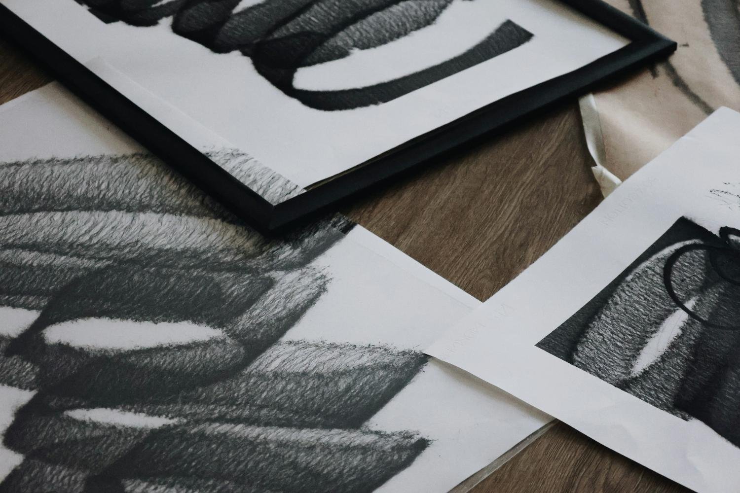 Close-up of black and white sketches and a framed artwork on a wooden surface, featuring abstract and geometric shapes.