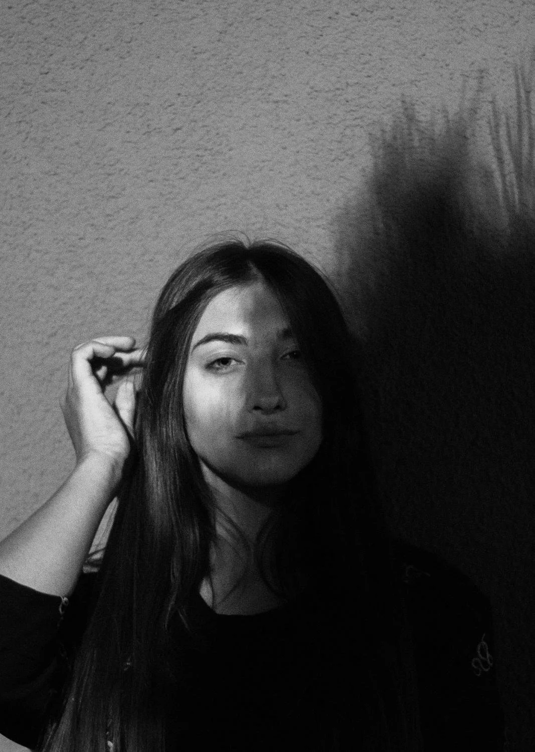 Black and white photo of a young woman with long hair, looking at the camera with a neutral expression, standing against a textured wall, with her hand near her ear, and a shadow cast on the wall.
