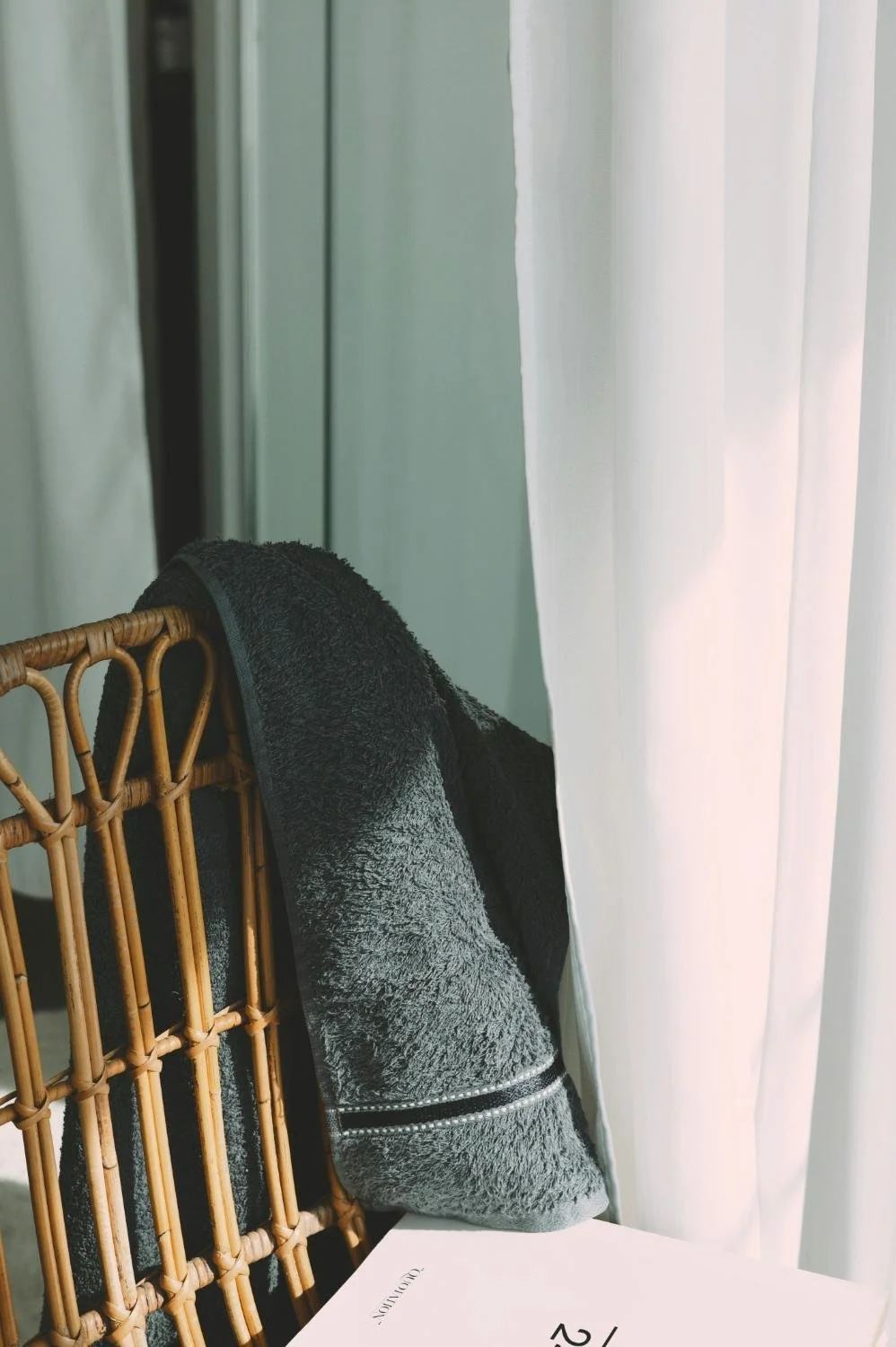 A corner of a wicker chair displaying a black and gray towel, with a curtain and part of a paper or magazine on a table in the background.