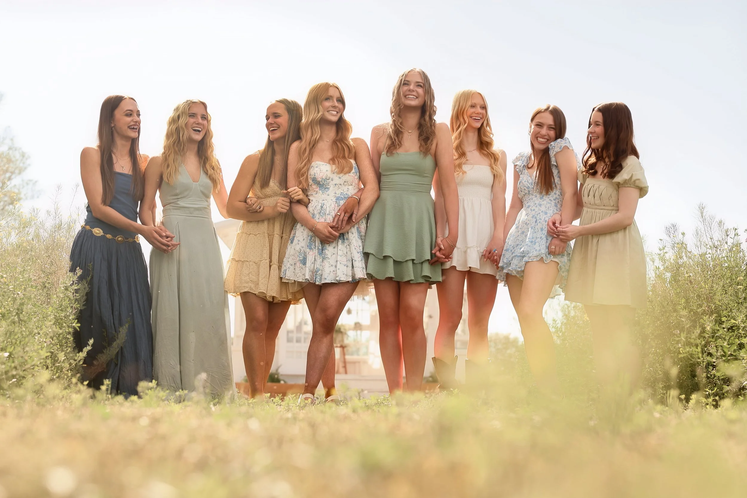A group of nine young women standing closely together outdoors, smiling and holding hands, dressed in casual summer clothing.