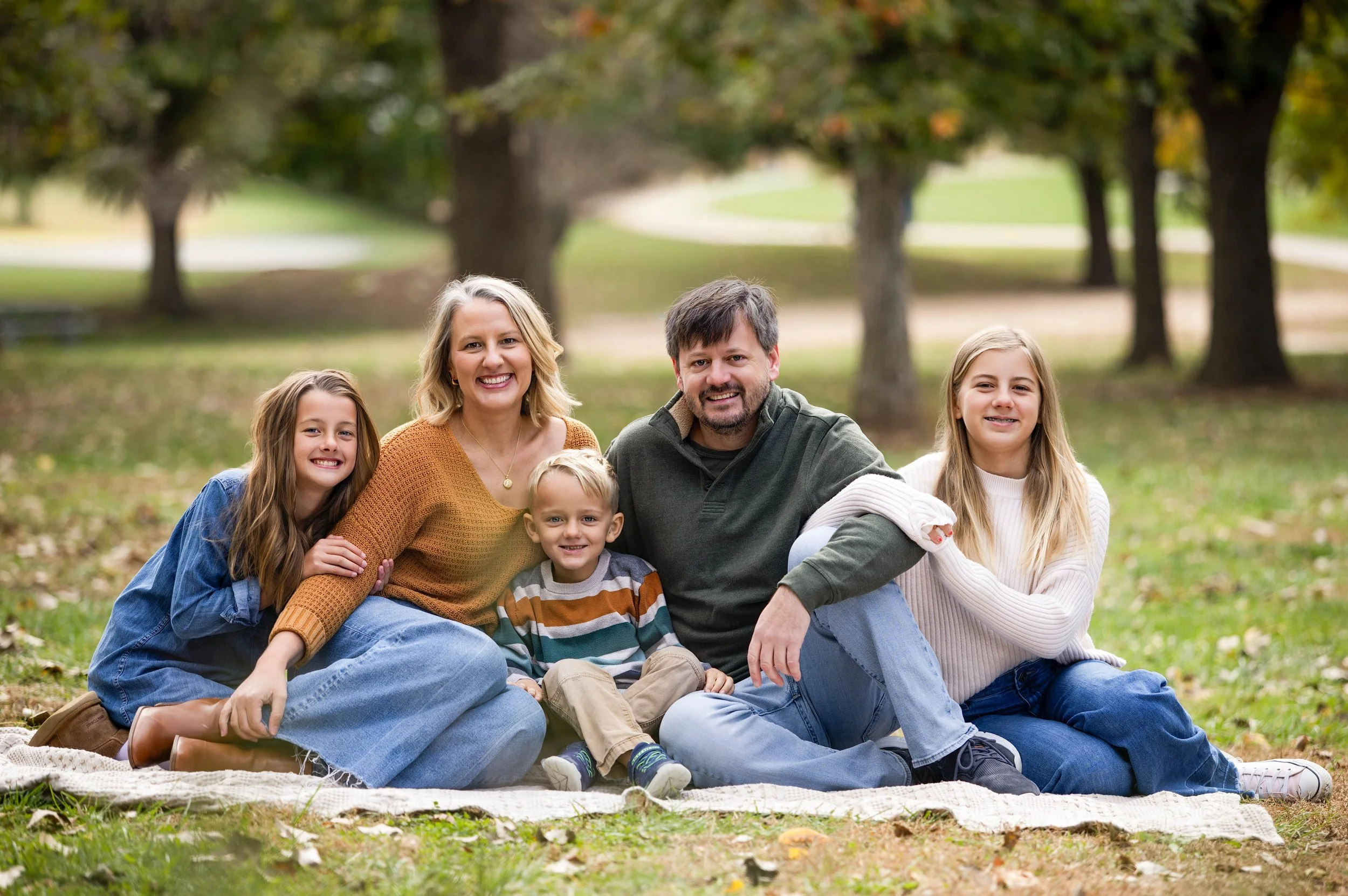 Family of six, including two young girls, two boys, a woman, and a man, sitting together on a blanket outdoors in a park with trees and grass in the background.
