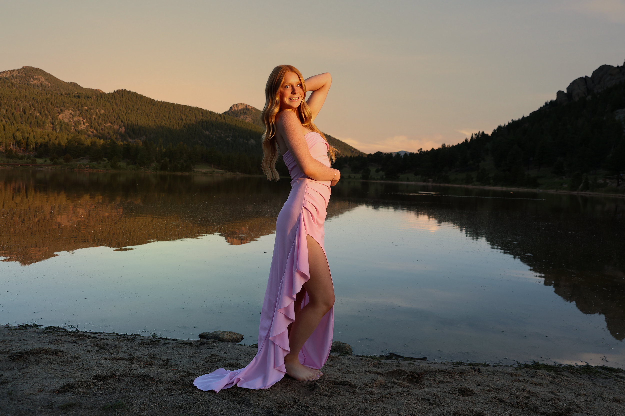 A woman in a pink dress standing barefoot by a lake at sunset, with mountains and trees in the background, smiling and posing with one hand behind her head.