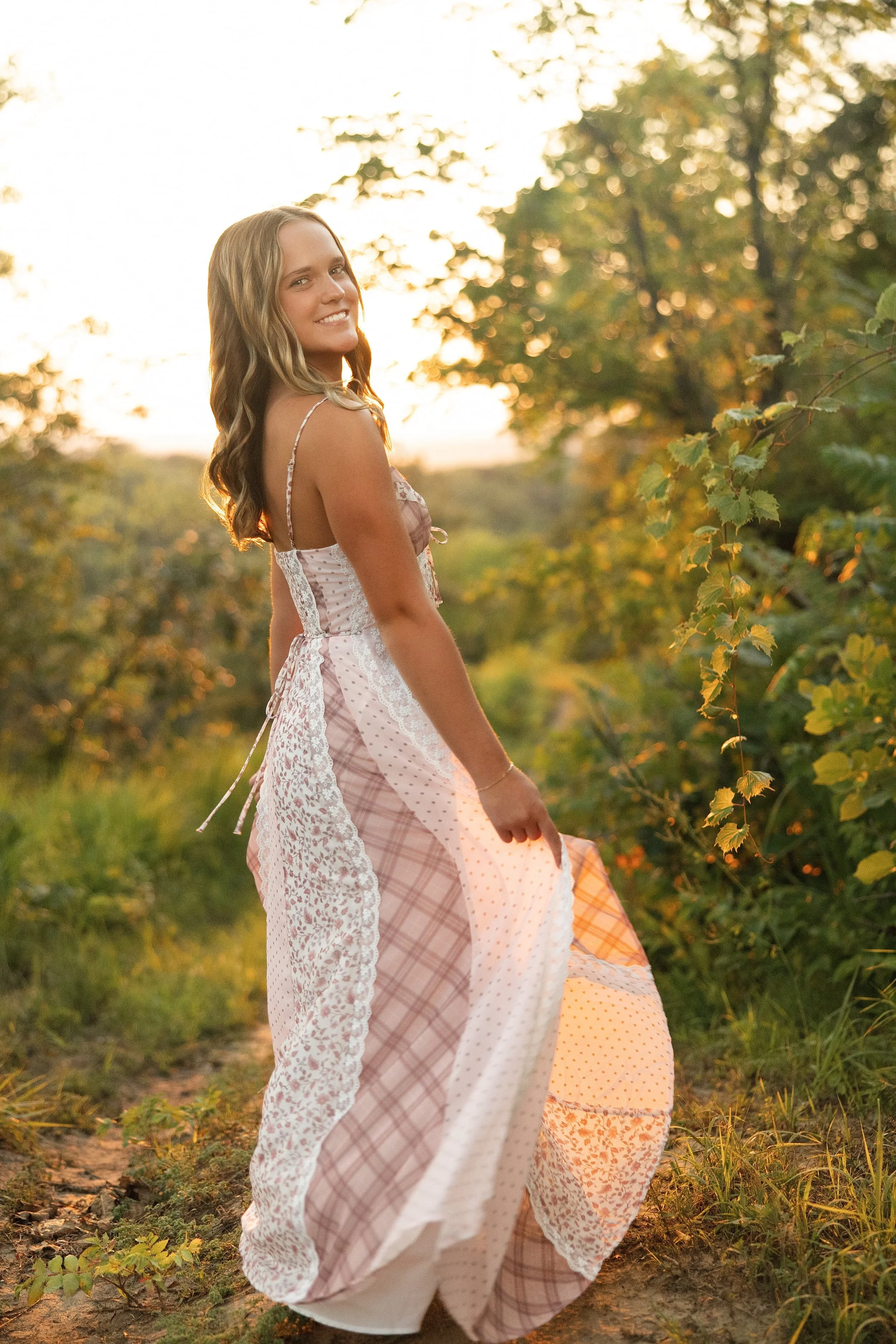 A smiling young woman with long wavy blonde hair wearing a light pink plaid and white lace dress walking through a vineyard during sunset.