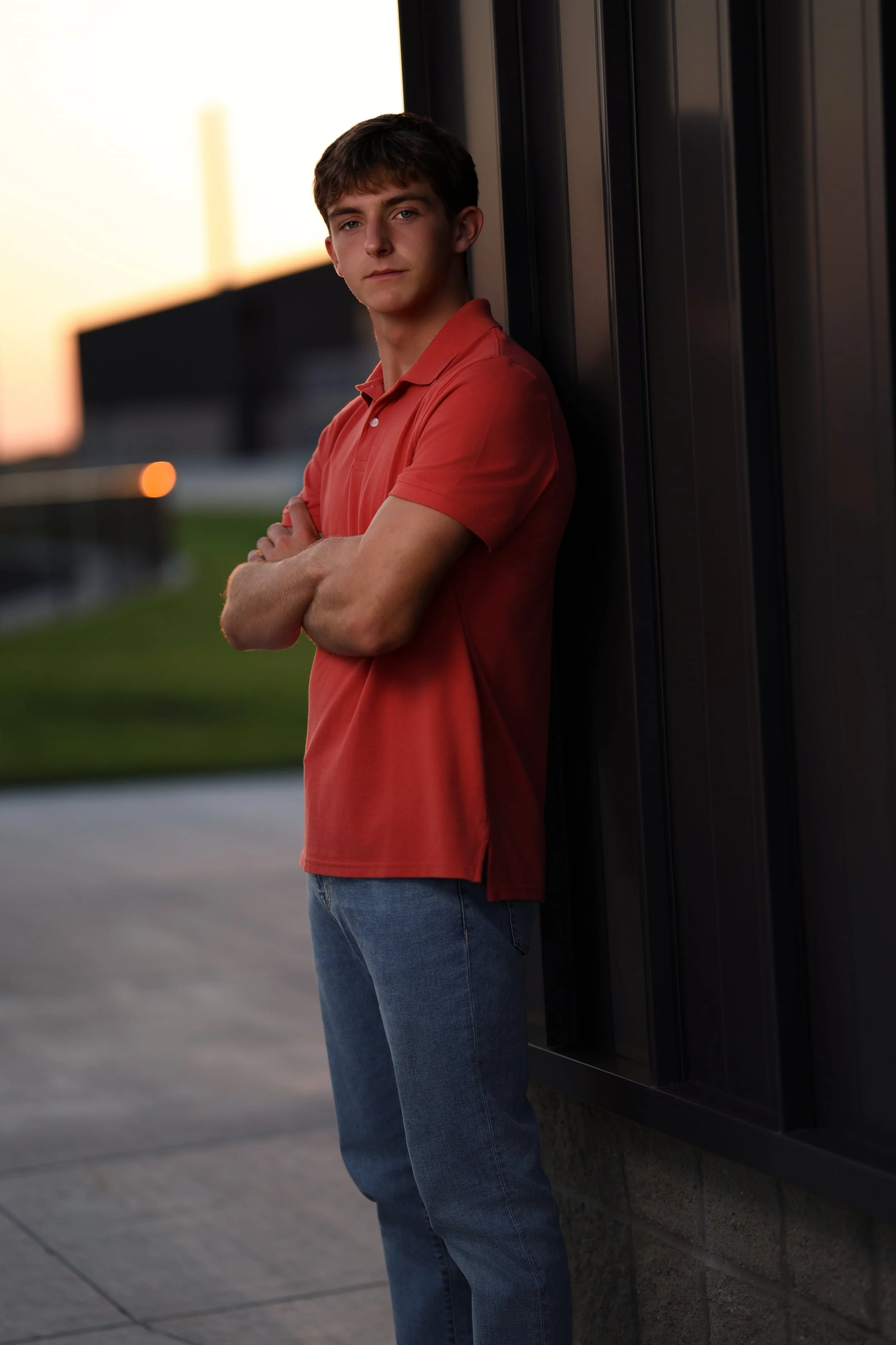 A young man wearing a red polo shirt and blue jeans leaning against a black metal wall outdoors during sunset, with arms crossed and looking at the camera.