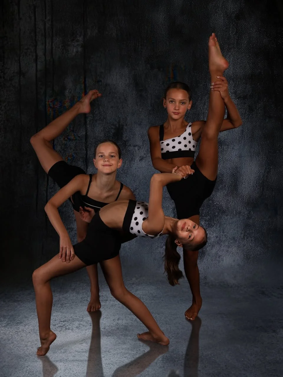 Three young girls performing gymnastic poses together against a dark background, wearing black and white polka dot leotards.
