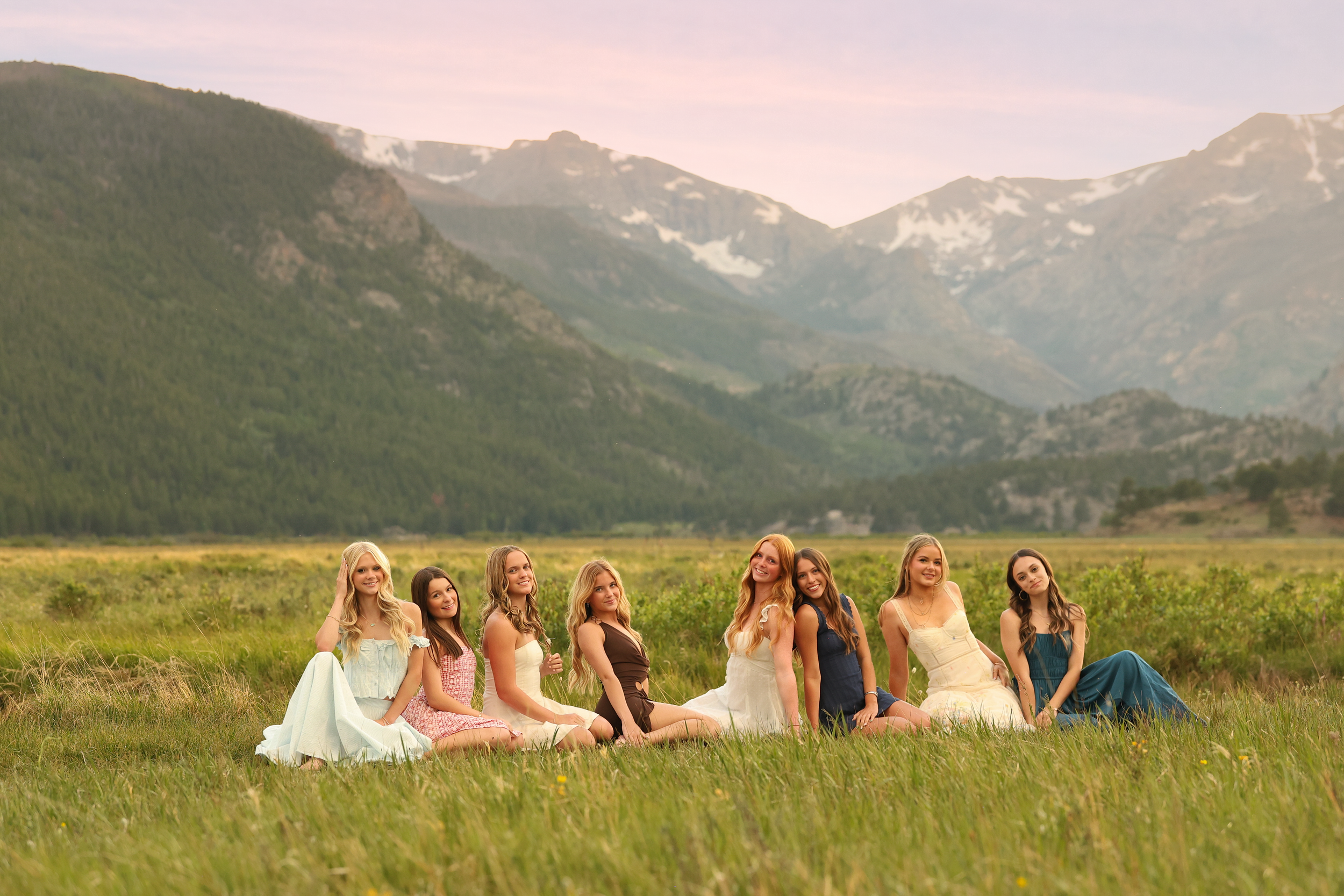 Group of young women in dresses sitting on grass in a meadow with mountains and cloudy sky in the background.
