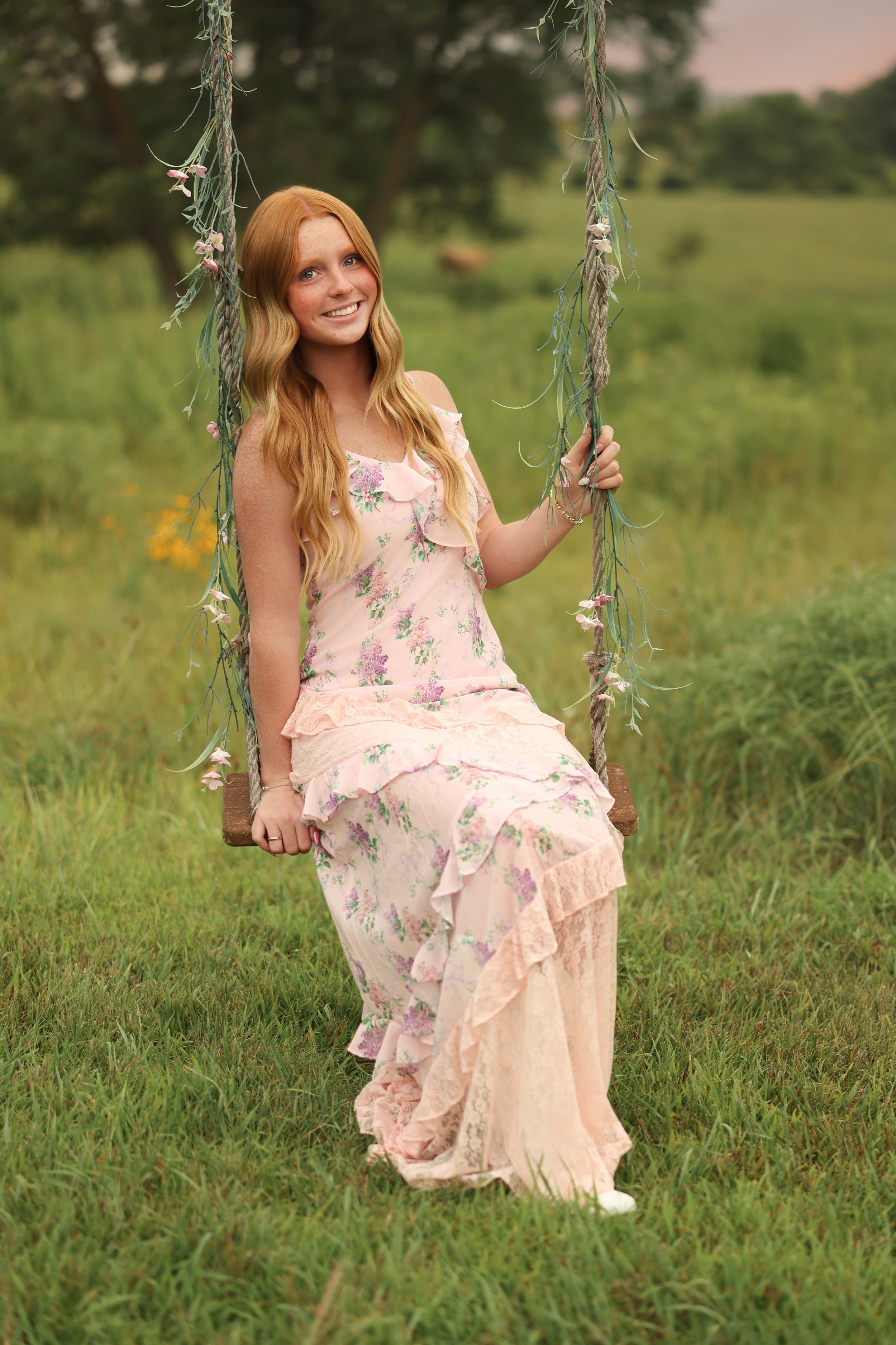 A young woman with long red hair, wearing a pastel floral dress, sitting on a swing outdoors in a grassy field with trees in the background, smiling at the camera.