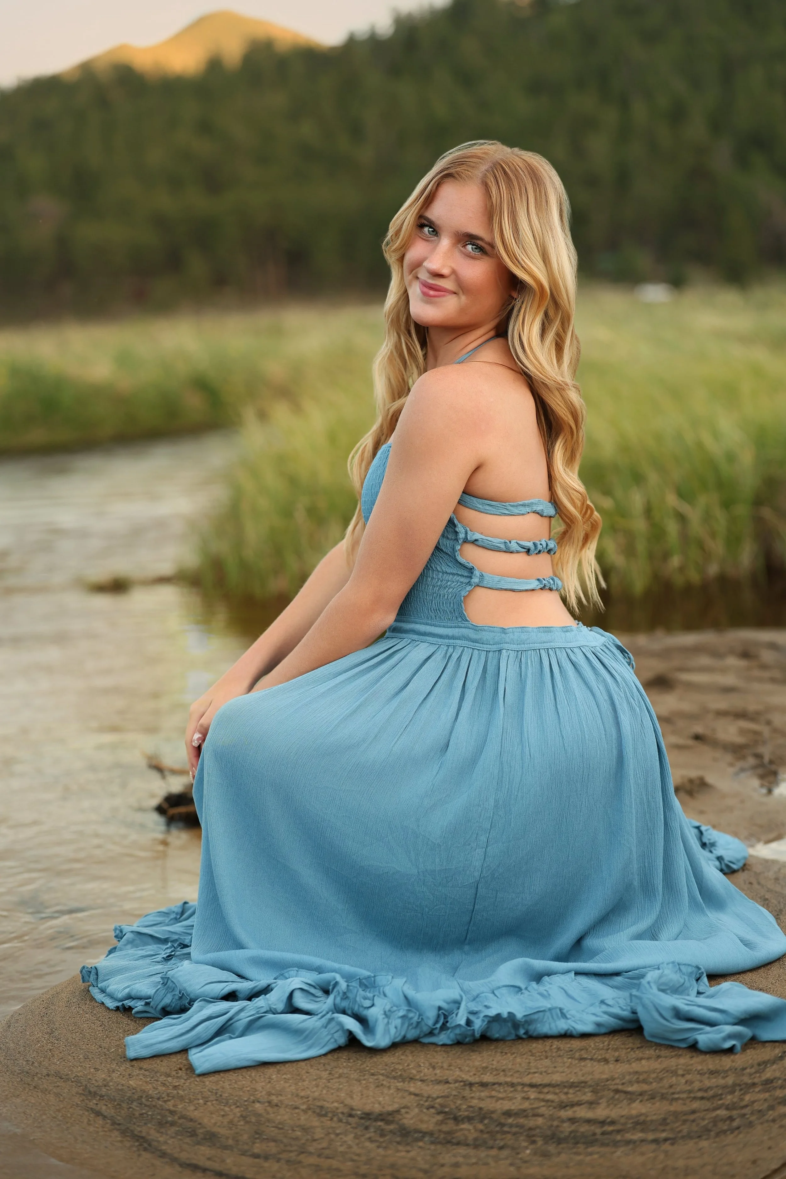 A young woman with long blonde hair sitting on a sandy beach near a body of water, wearing a flowing blue dress with cut-out details on the back, smiling softly, with a blurred background of trees and mountains.