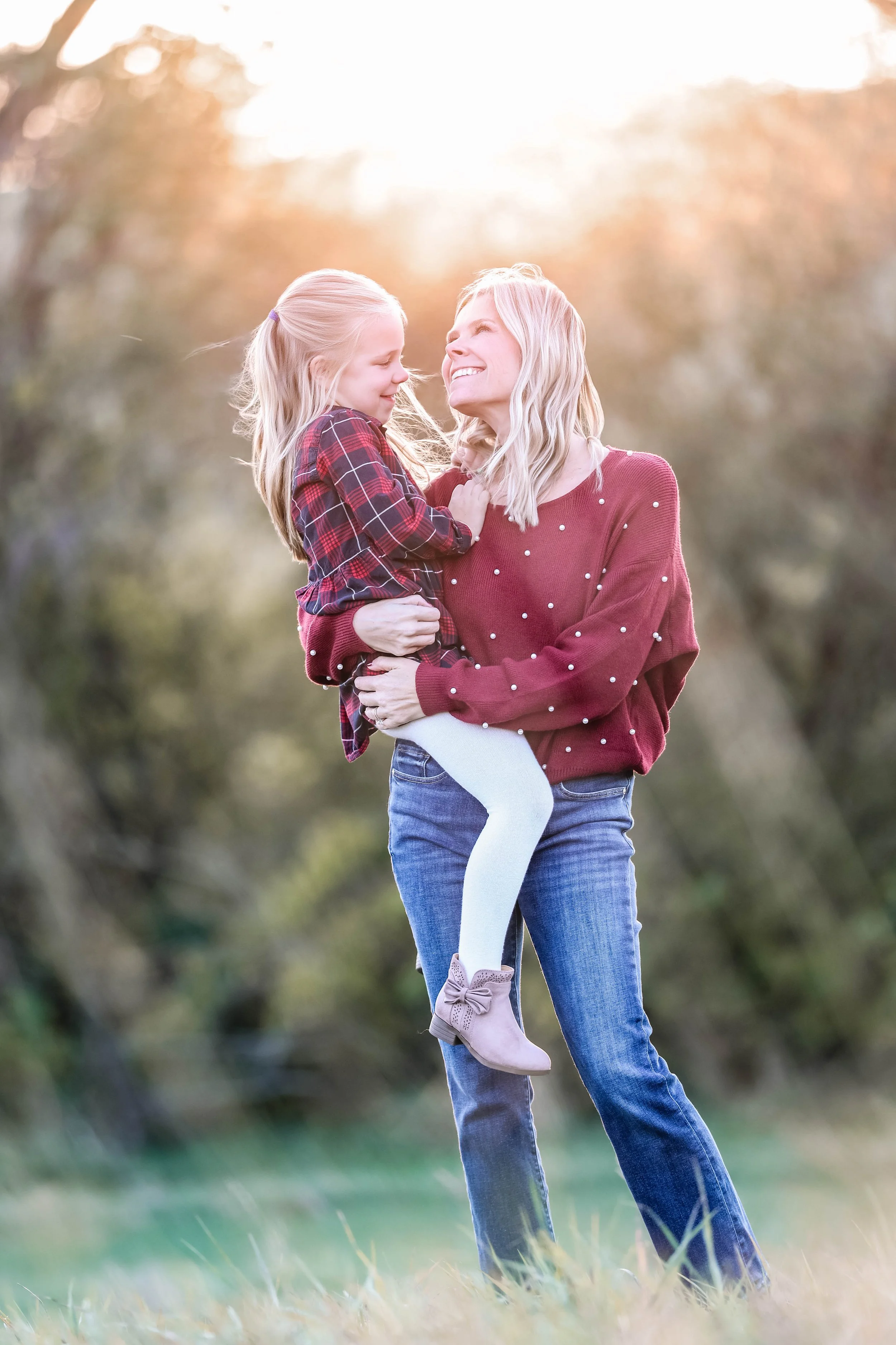 A woman holding a young girl in her arms outdoors during sunset, both smiling and looking at each other. The woman is wearing a red sweater with white polka dots and blue jeans, while the girl is wearing a plaid shirt and white leggings.