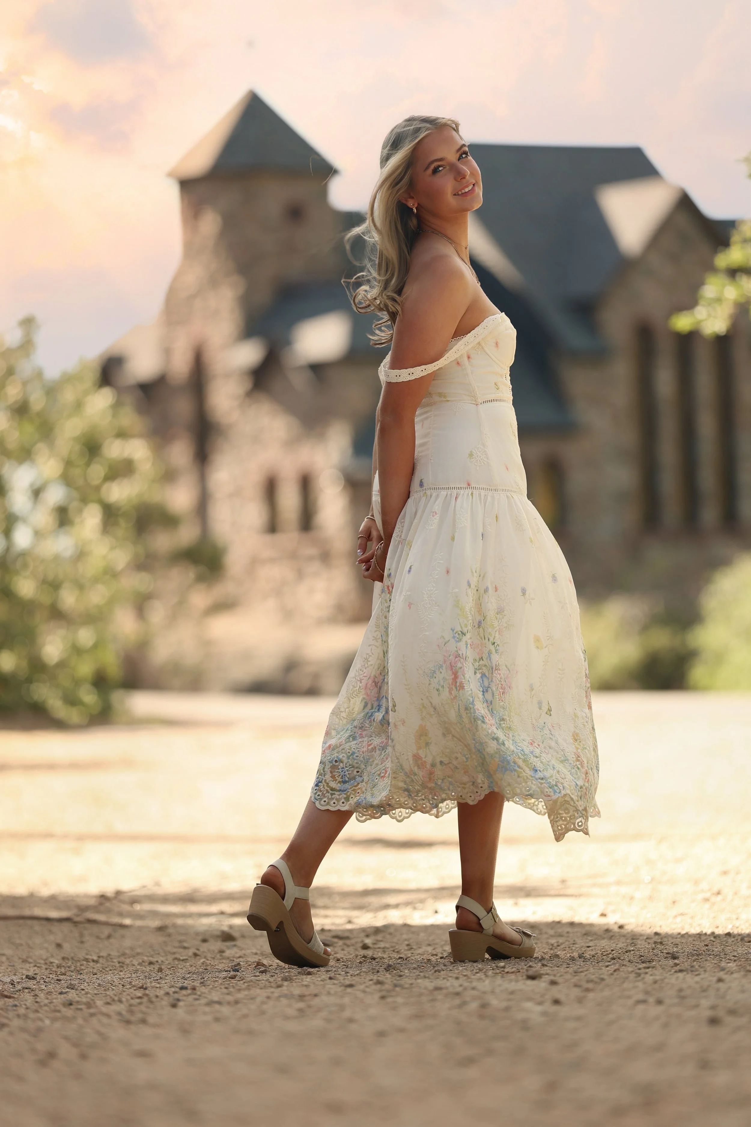 A woman in a white dress with floral patterns and lace trim is standing outdoors on a dirt path. She is smiling and looking at the camera, with a blurred historic stone building in the background during the sunset.
