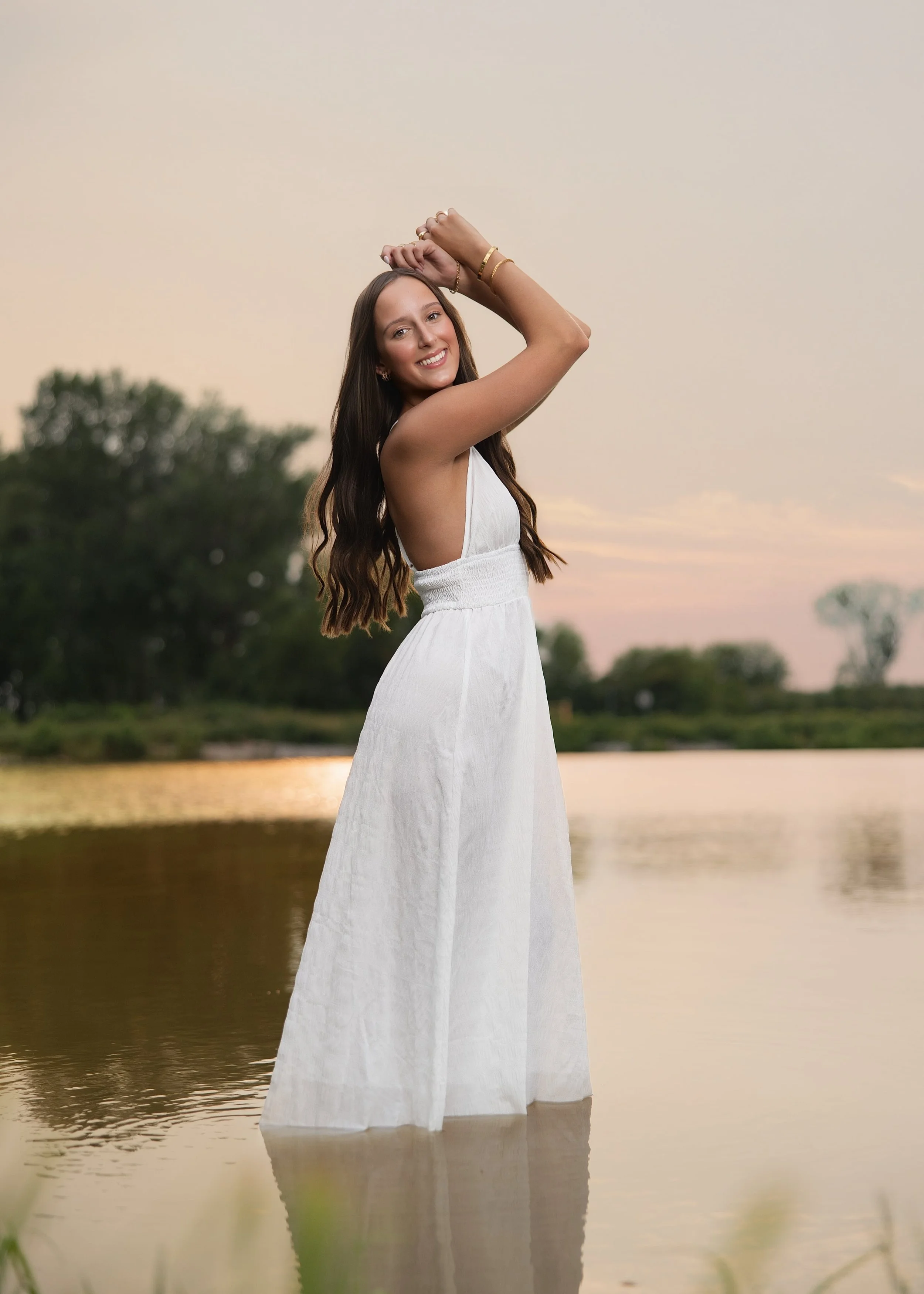A woman with long dark hair wearing a white dress, smiling and standing in a shallow body of water during sunset, with trees blurred in the background.