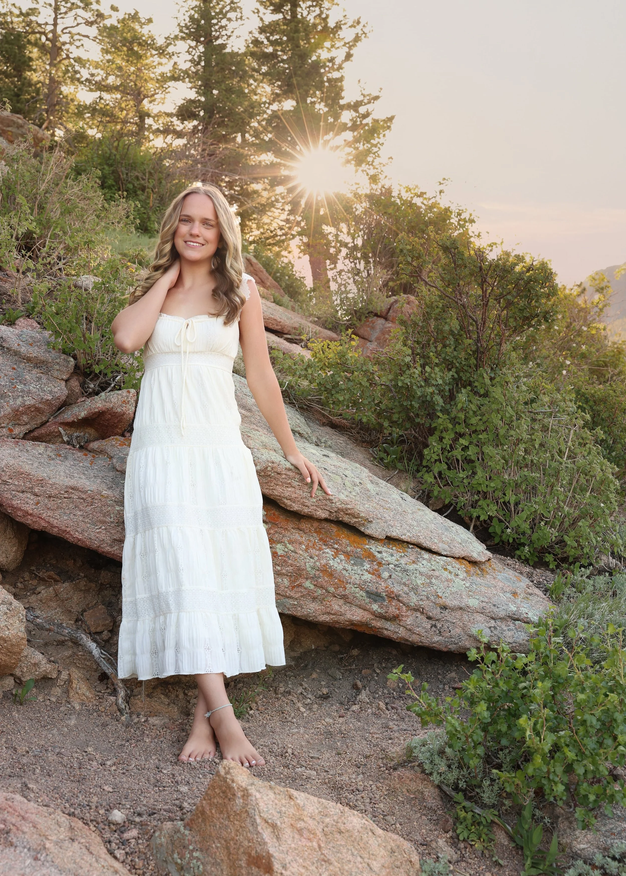 A young woman in a white dress standing among rocks and greenery, with sunlight shining through trees in the background.