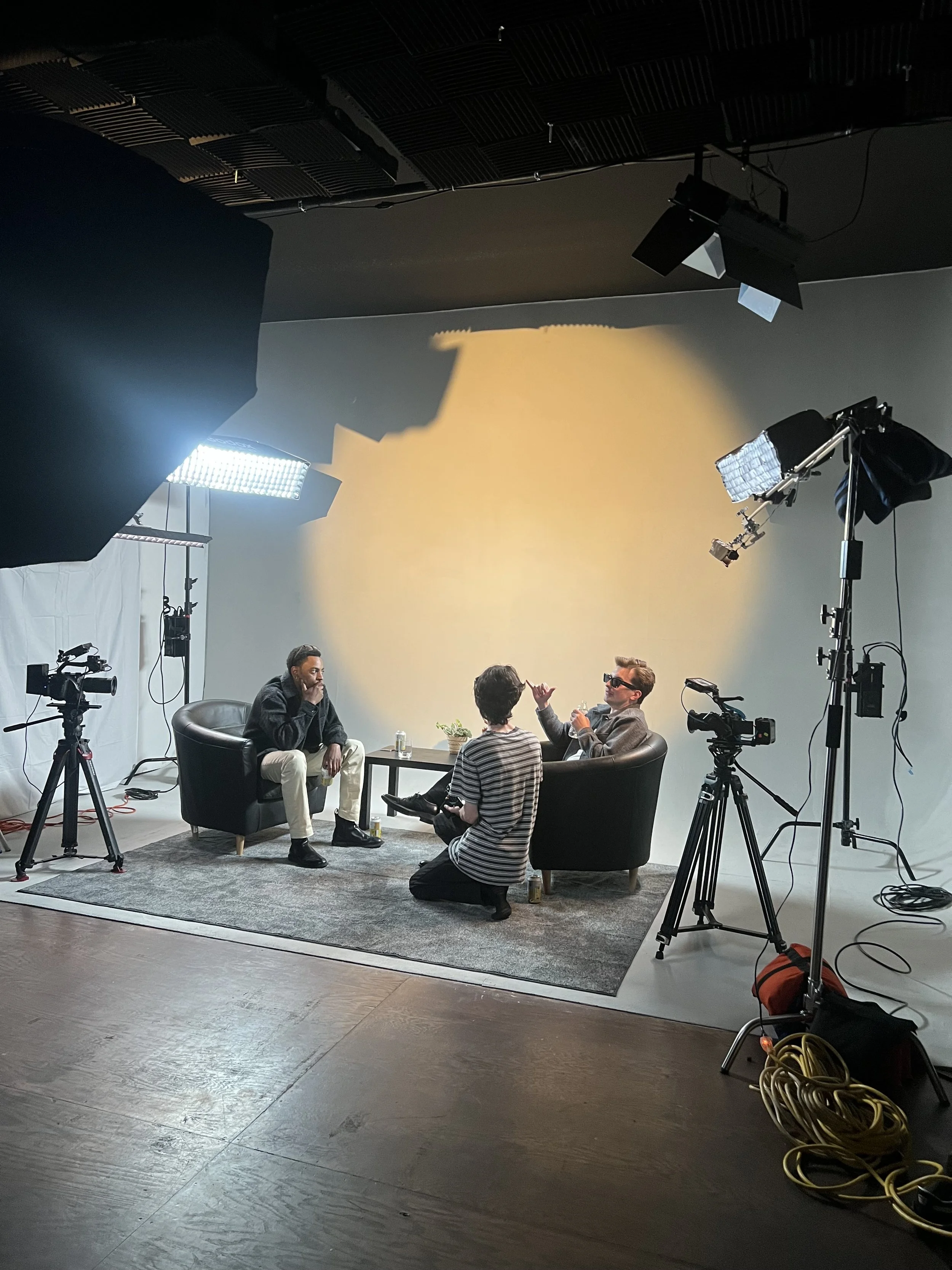 A behind-the-scenes view of a video or photo shoot studio with two men seated on black chairs, a man kneels on the floor nearby, a director and crew member, with professional lighting equipment, cameras, and other filming gear surrounding them.