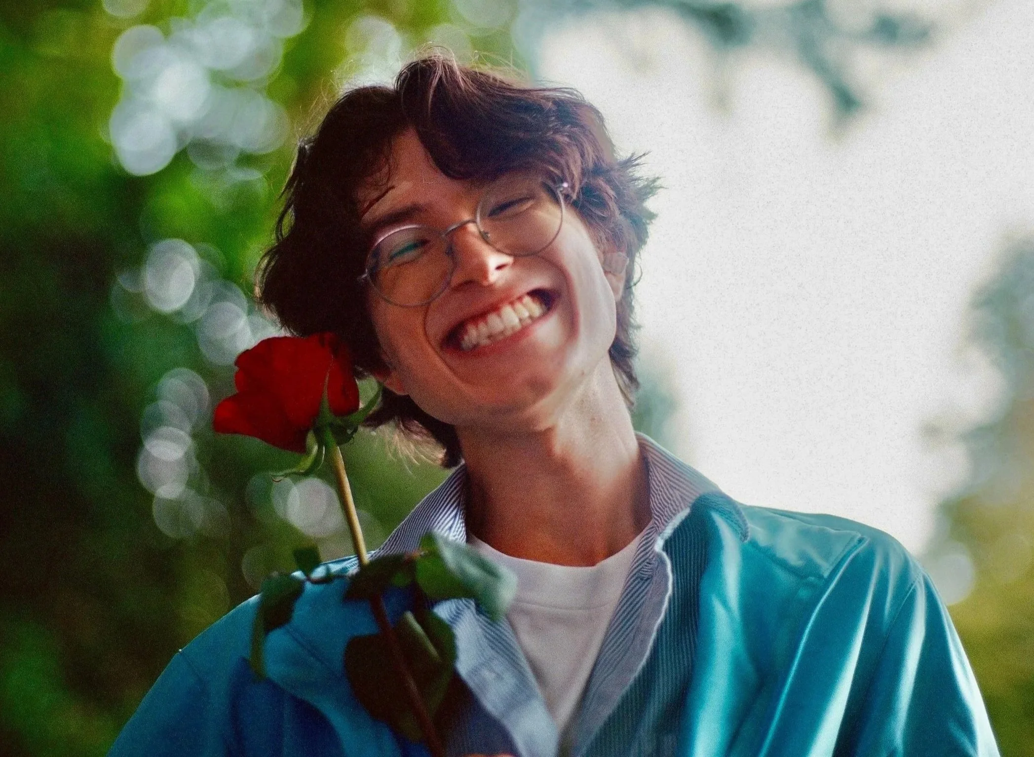 A young man with glasses and curly hair smiling broadly while holding a red rose. Blurred greenery in the background.