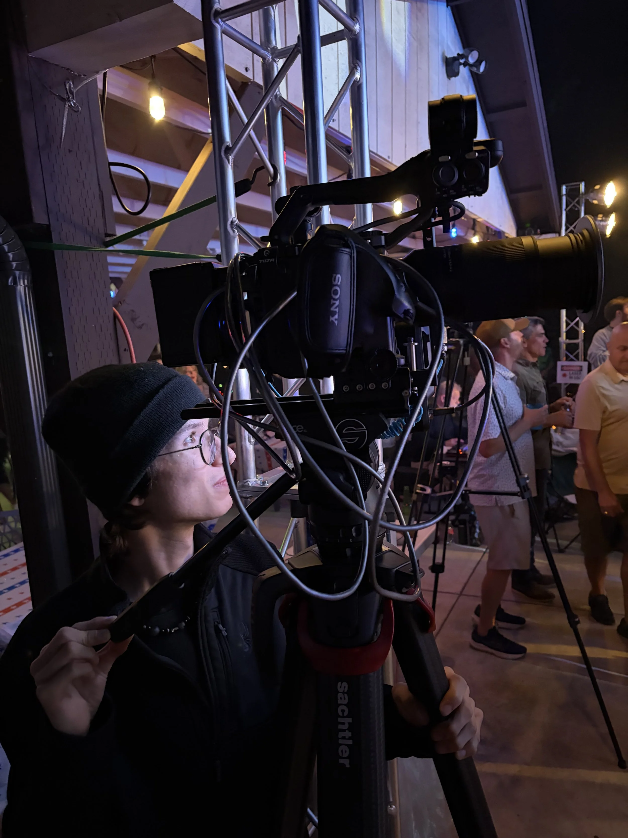 A man operating a professional camera using a tripod at an outdoor event with people in the background.