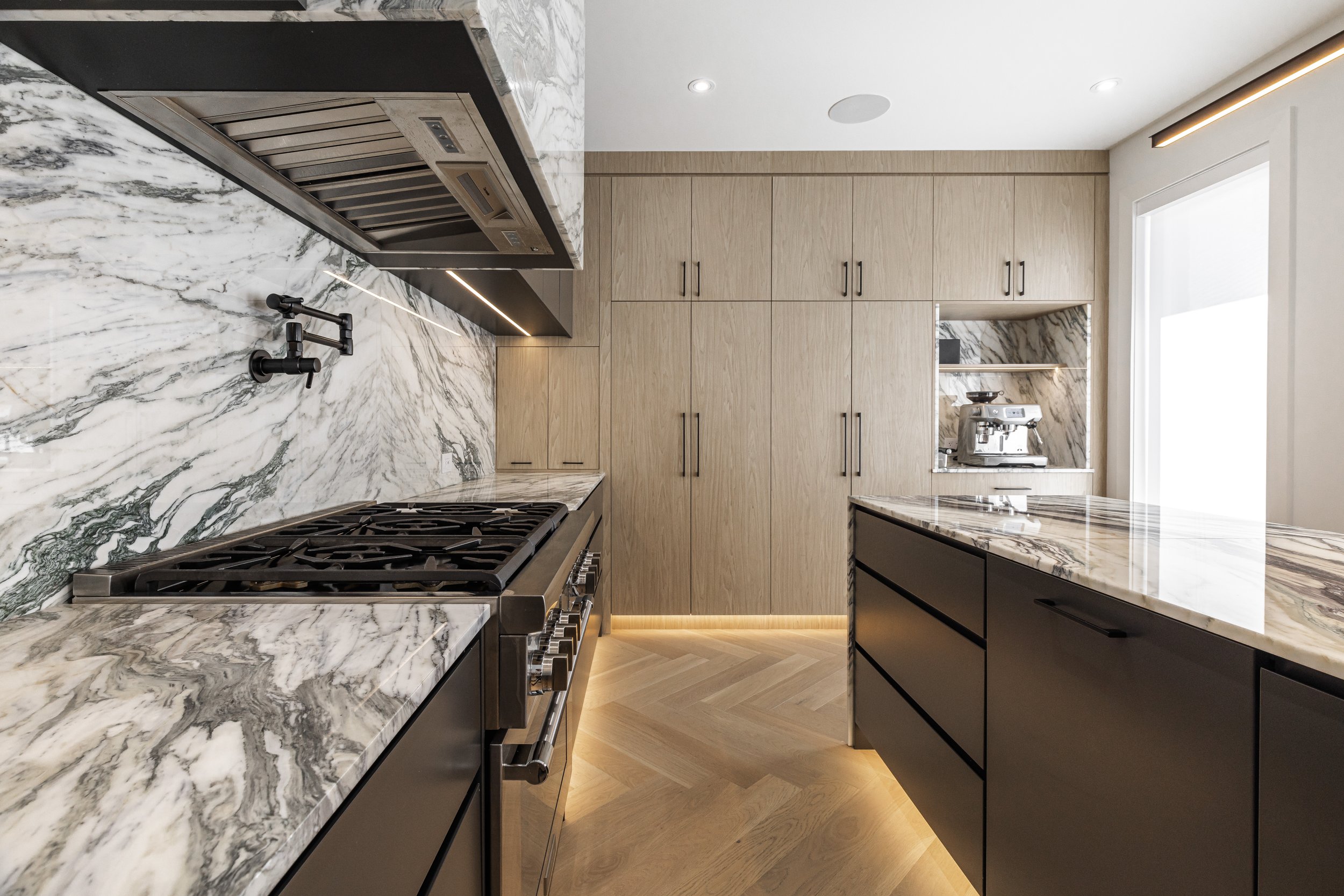 Modern kitchen with marble countertops and backsplash, black cabinetry, built-in oven, and a coffee machine on the nook. Light wood cabinets and flooring with integrated lighting.