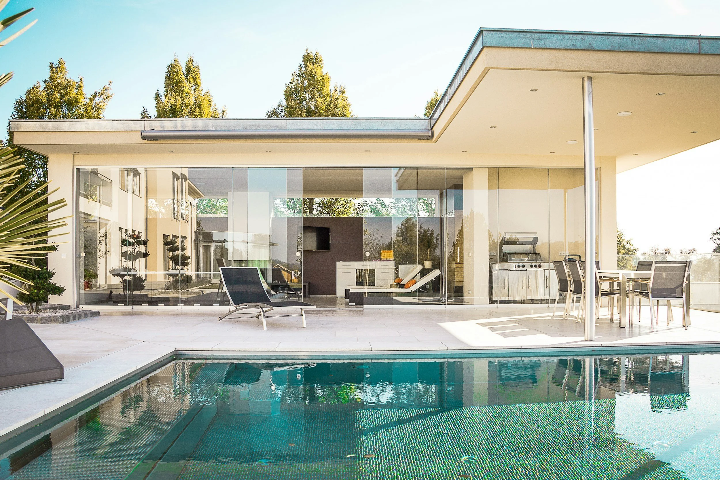 Modern house with a glass wall, outdoor pool, lounge chairs, patio dining set, and barbecue grill, surrounded by trees.