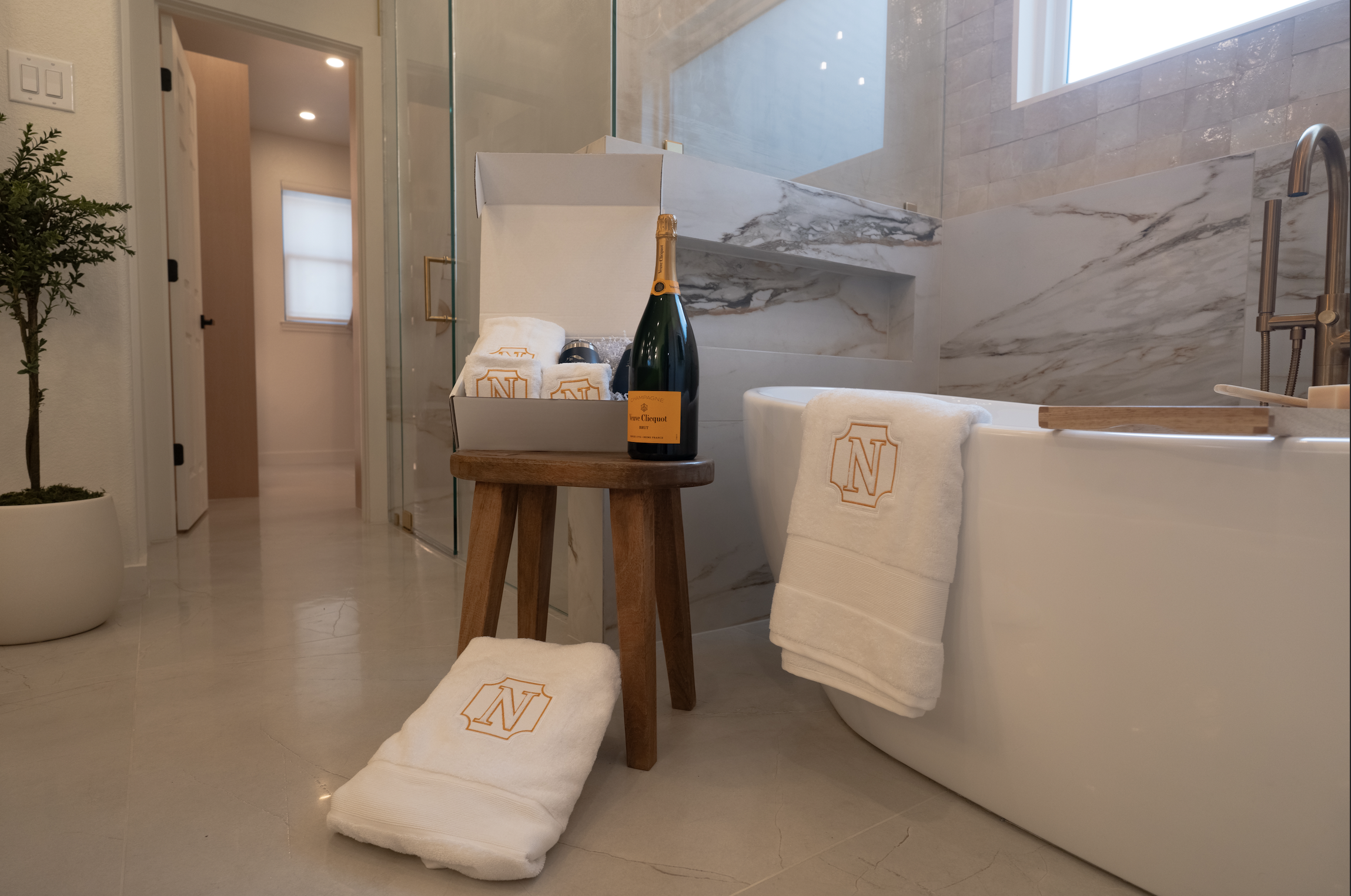 Bathroom with a bathtub, a small wooden stool with a bottle and towels, a towel on the bathtub with an embroidered letter 'N', and a rolled towel on the floor with the same embroidery. There is a window, a glass shower enclosure, and a potted plant i