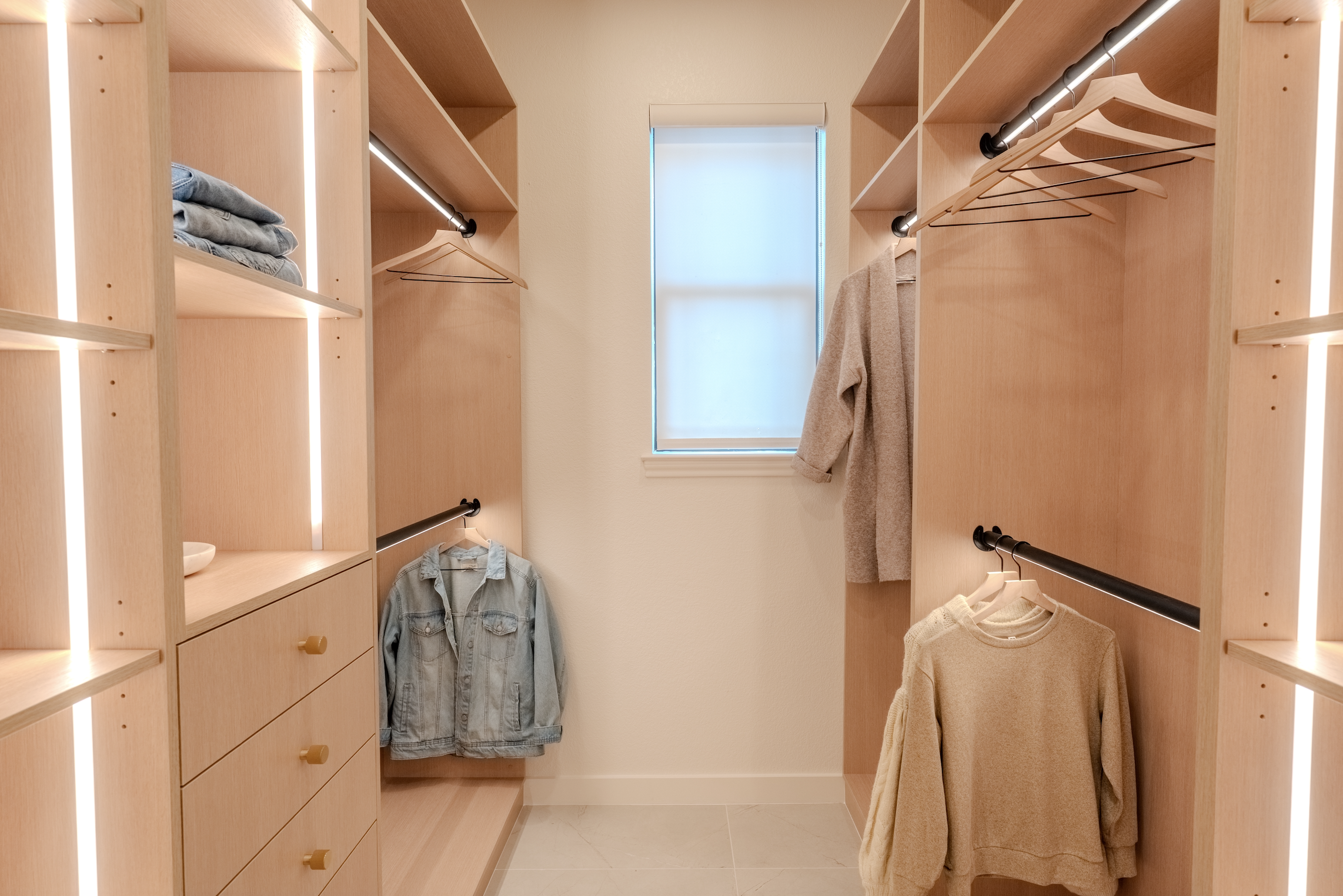 Empty walk-in closet with wooden shelves, hanging rods, a small window with a shade, and a few clothes including jackets and sweaters.