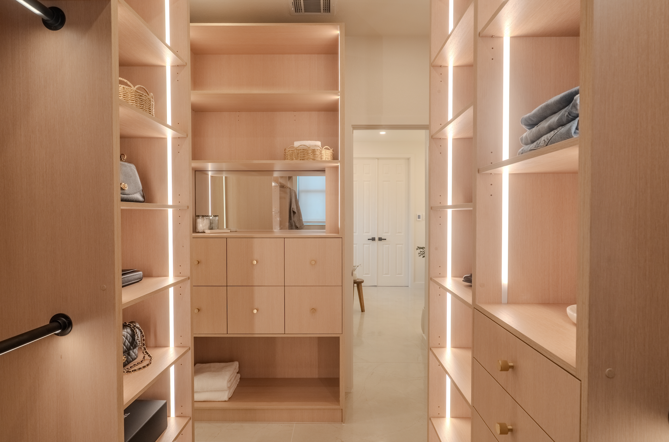 A walk-in closet with light wood shelves, drawers, and cabinets, containing folded towels, handbags, and other accessories, illuminated by vertical LED strip lights.