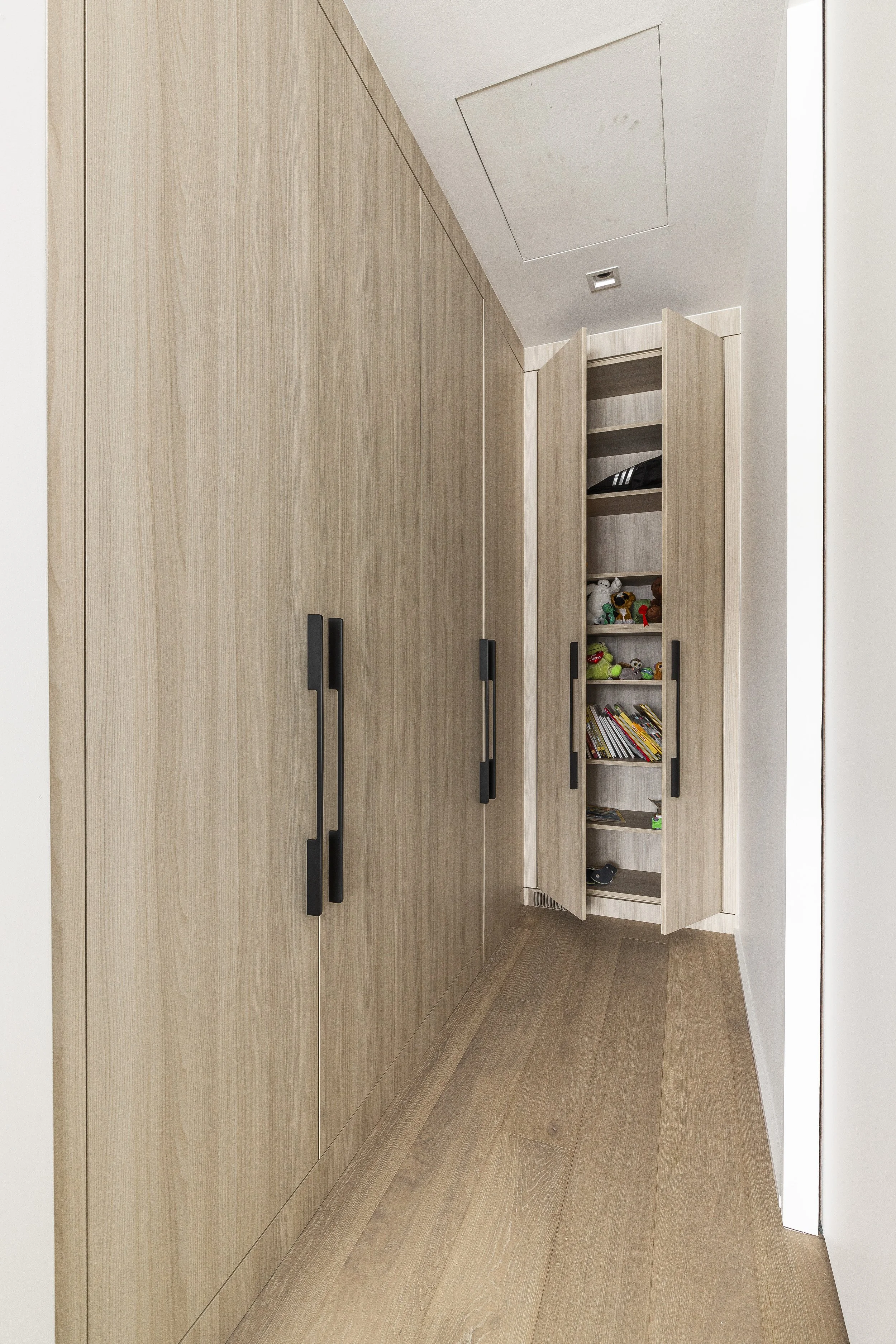 A hallway with wooden closet doors and a small open cabinet with shelves holding children's toys, books, and miscellaneous items.