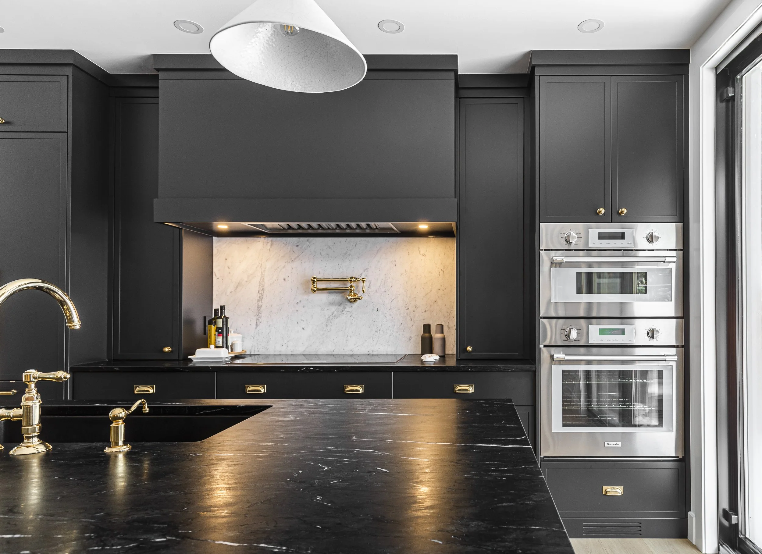 Modern kitchen with black cabinetry, marble backsplash, stainless steel oven, and gold fixtures.