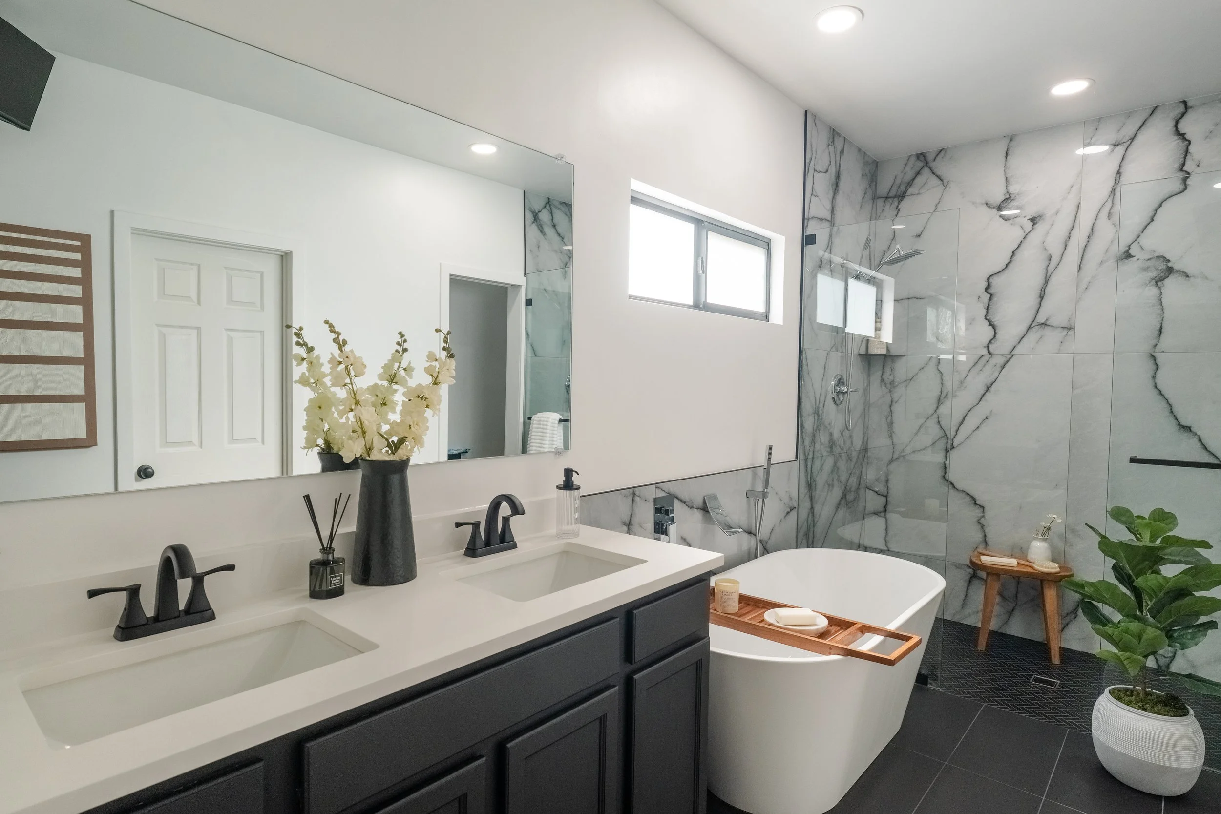Modern bathroom with a double vanity, black cabinets, white countertop, large mirror, and black fixtures. There is a standalone bathtub with a wooden tray, a walk-in shower with marble tiles, a small wooden stool, a potted plant, and a window letting