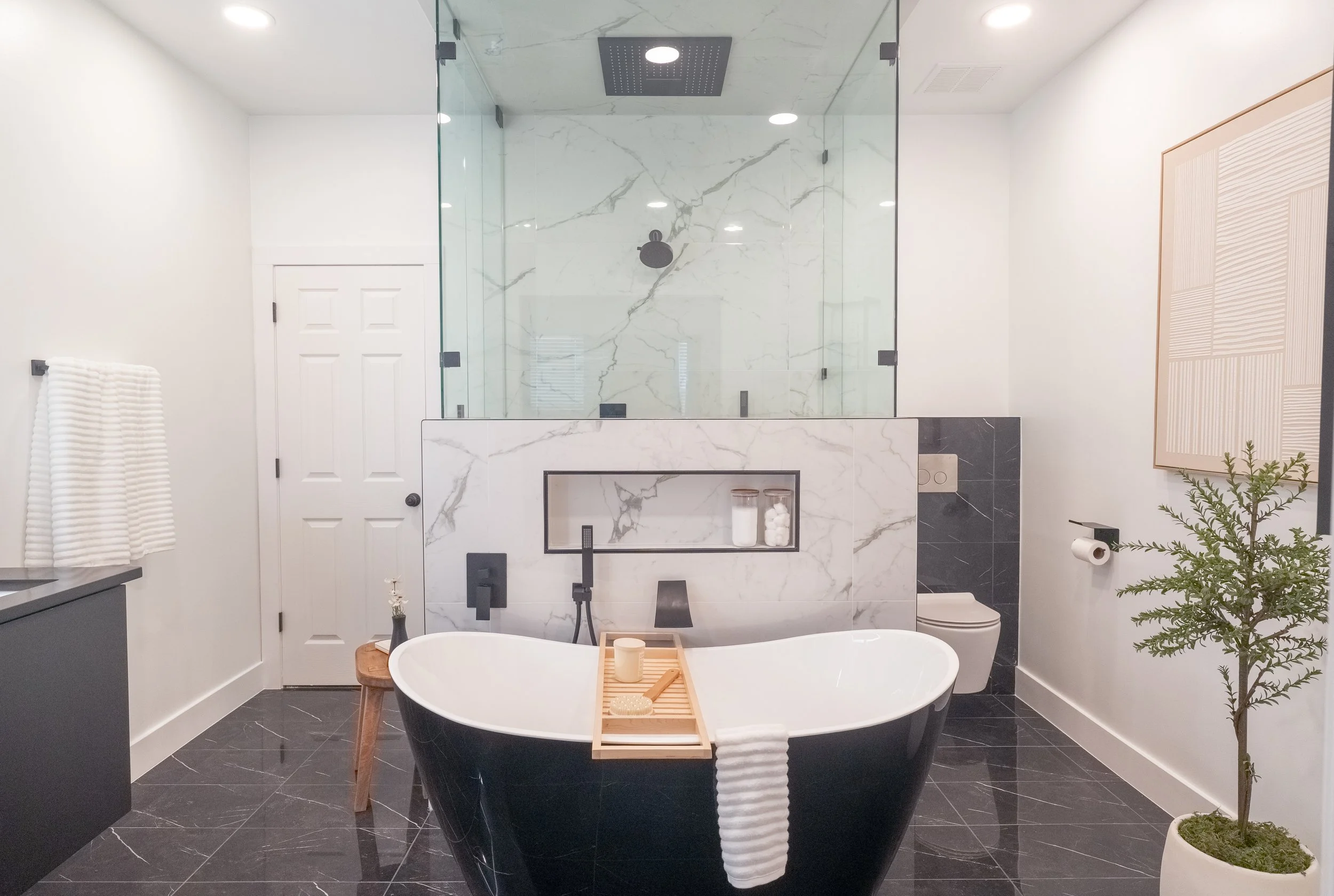 Modern bathroom with a freestanding black and white bathtub, a glass-enclosed shower with marble walls, a small wooden stool, a potted plant, and minimalist decor.