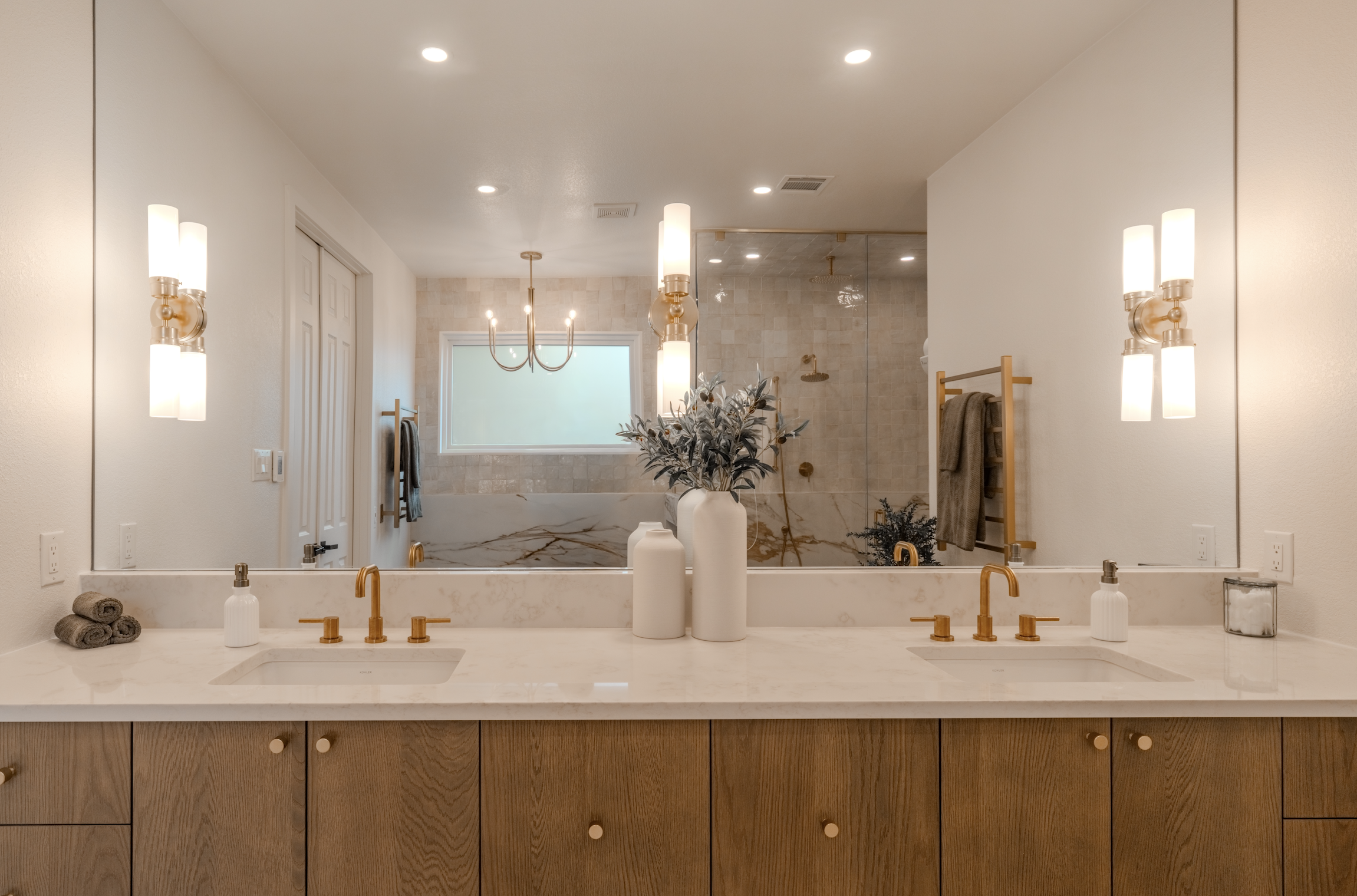 Modern bathroom with dual sinks and large mirror, featuring gold fixtures, a marble countertop, decorative vases, and a walk-in shower with a glass door and gold fixtures.