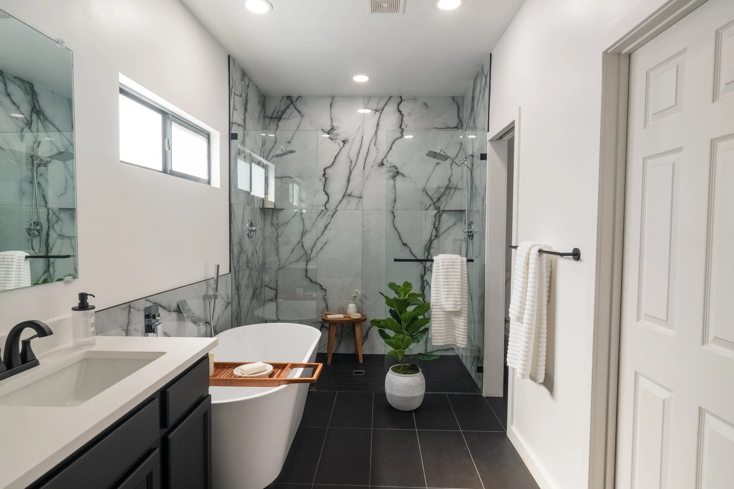 Modern bathroom with white walls, black floor tiles, a bathtub, double shower with marble walls, a mirror, a plant in a pot, and towels hanging on black towel bars.