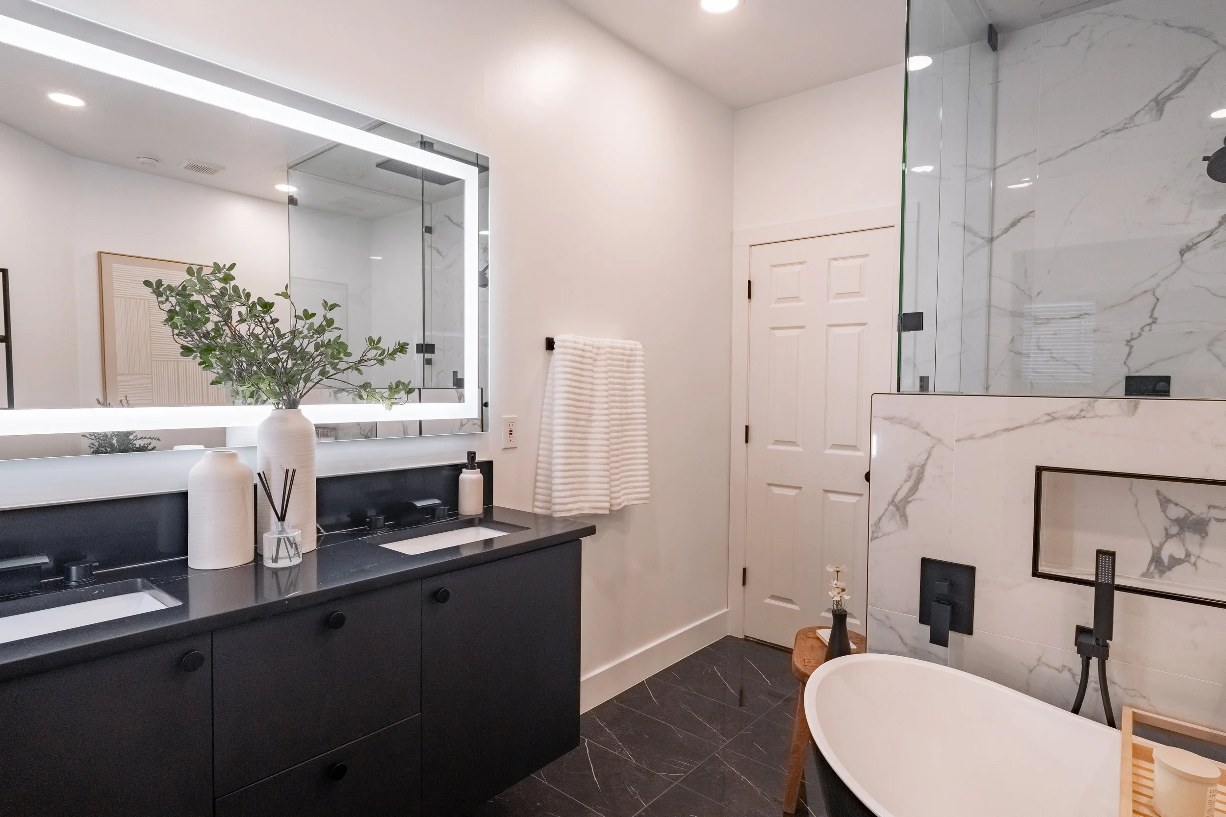 Modern bathroom with black vanity, white sink, large mirror with backlighting, glass-enclosed shower with marble walls, freestanding bathtub with wooden stool, white door, and decorative vases with greenery.