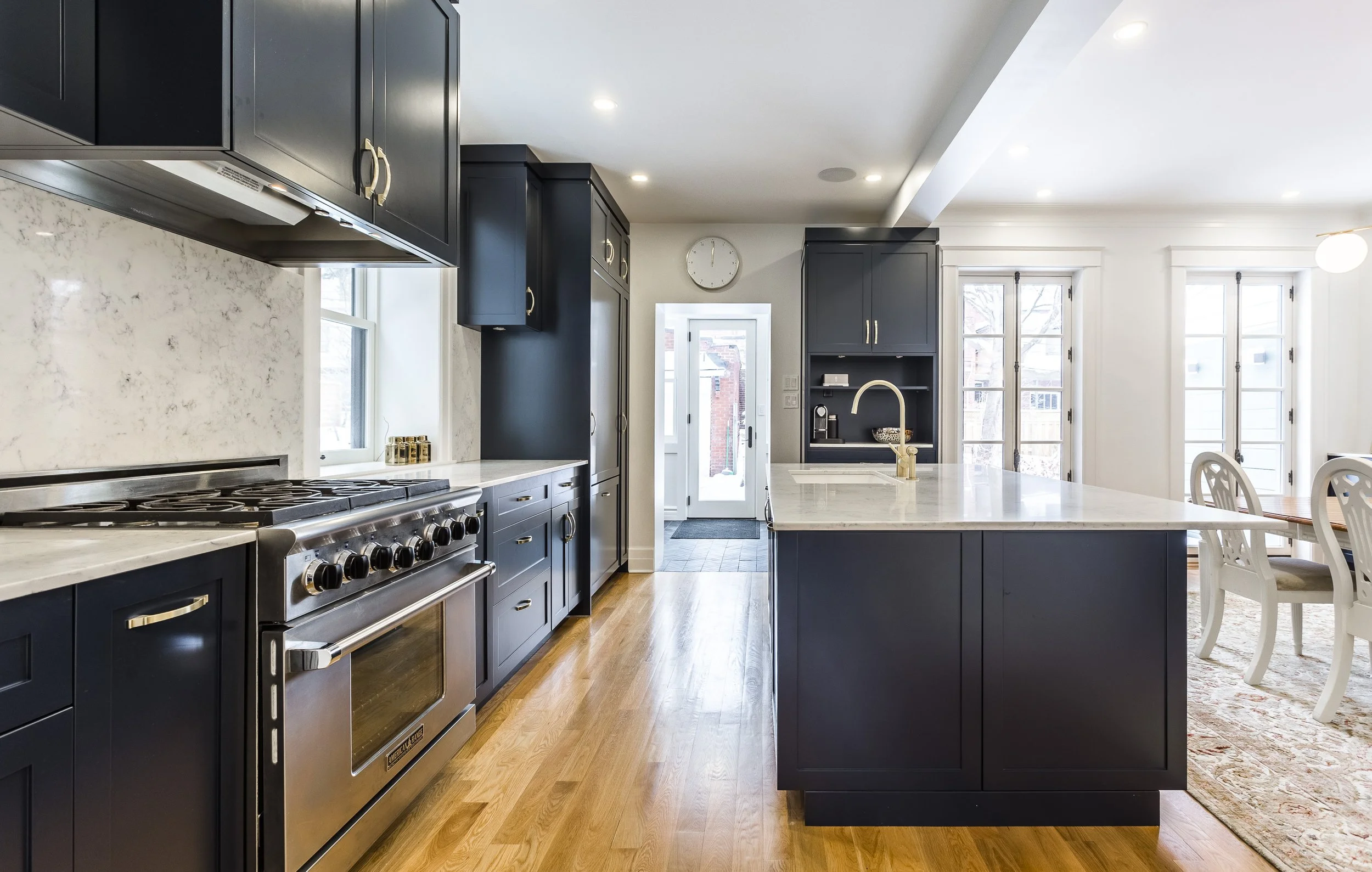 Modern kitchen with navy blue cabinets, a large kitchen island with a white countertop, stainless steel oven, and hardwood floors. Bright sunlight streams through multiple windows and glass doors.