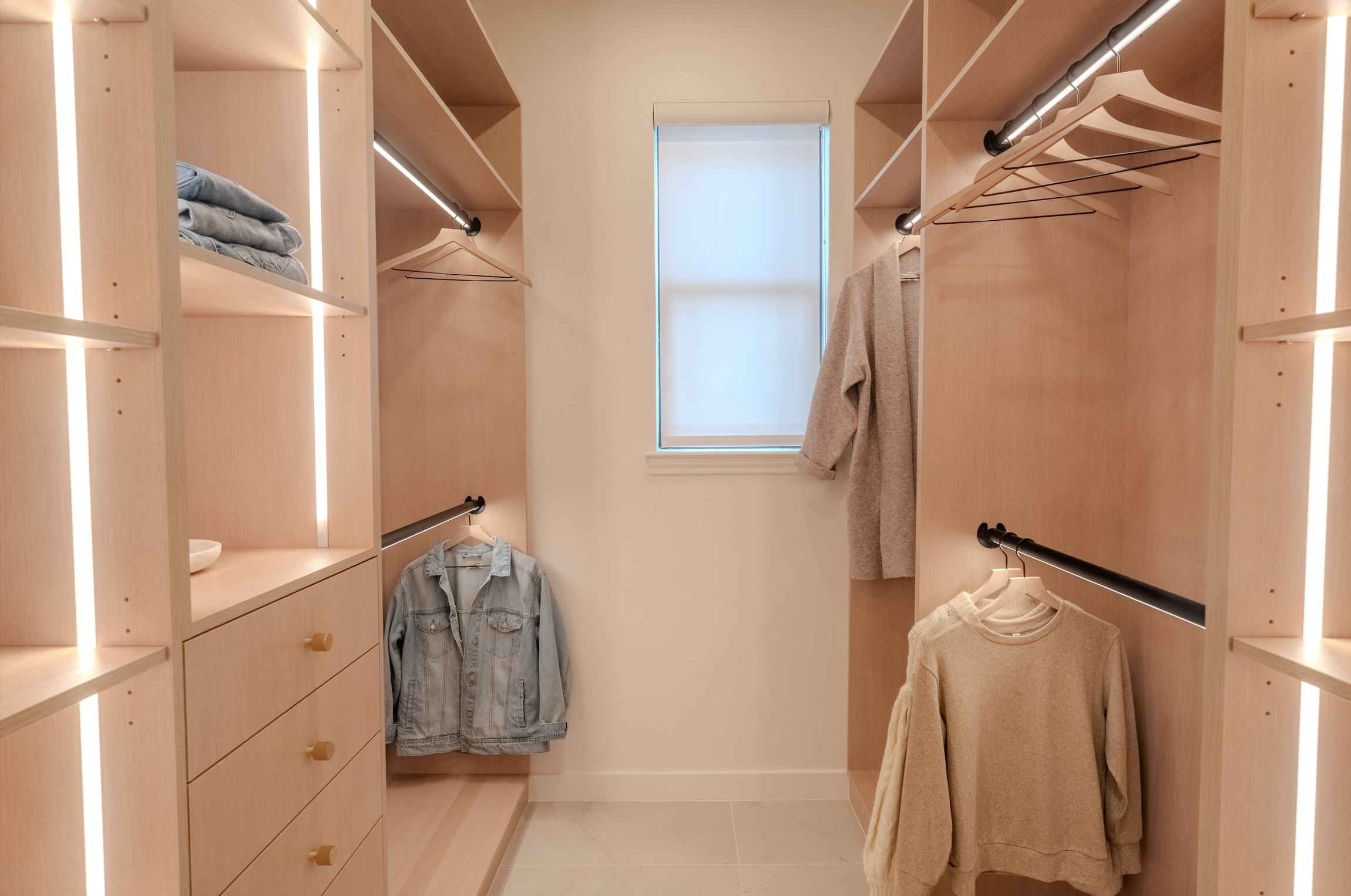 Walk-in closet with light wood shelving and hanging rods, some clothes and hangers, a small window at the end, and beige tiled floor.