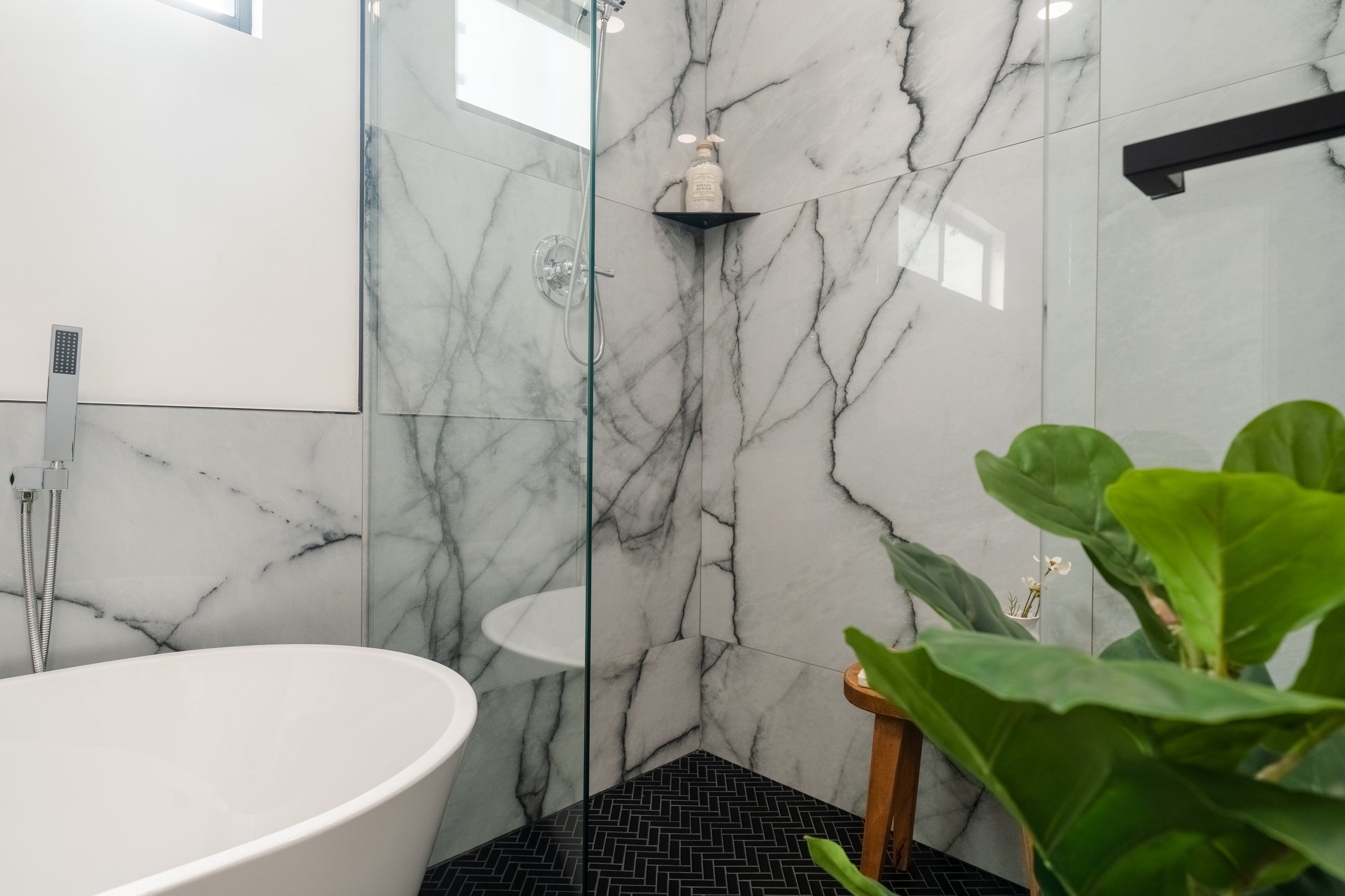 A modern bathroom featuring a walk-in shower with marble walls, a glass door, a black shelf holding a soap dispenser, a black shelf, a white freestanding bathtub, a black patterned floor, and a large green plant.
