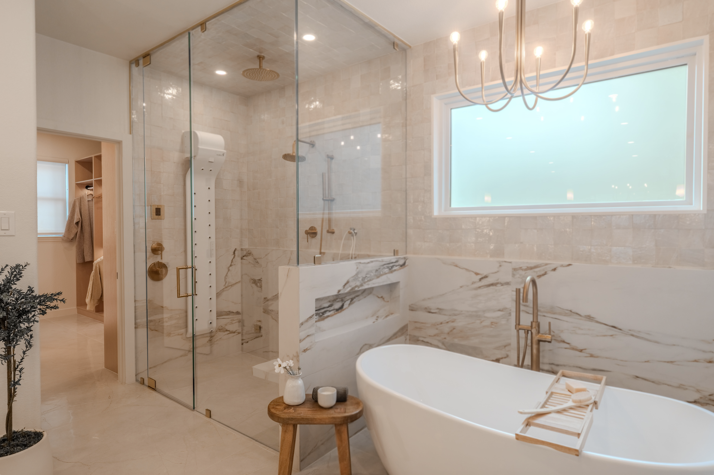 Modern bathroom featuring a freestanding white bathtub with a wooden tray, marble accents, a glass walk-in shower with multiple showerheads, a large frosted window, a chandelier, and a small side table with a vase and candles.