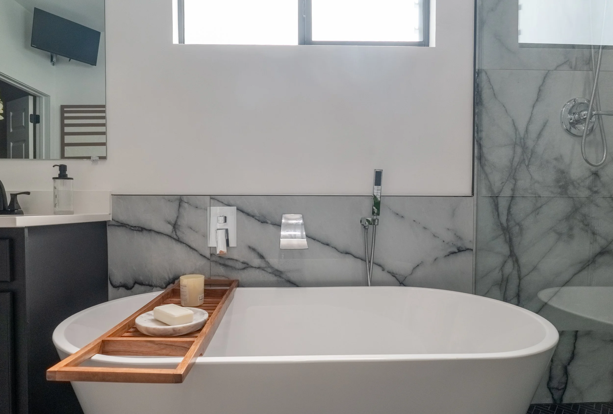 Modern bathroom featuring a white bathtub with a wooden tray holding soap and a candle, gray marbled walls, a showerhead, a mirror, and a window letting in natural light.
