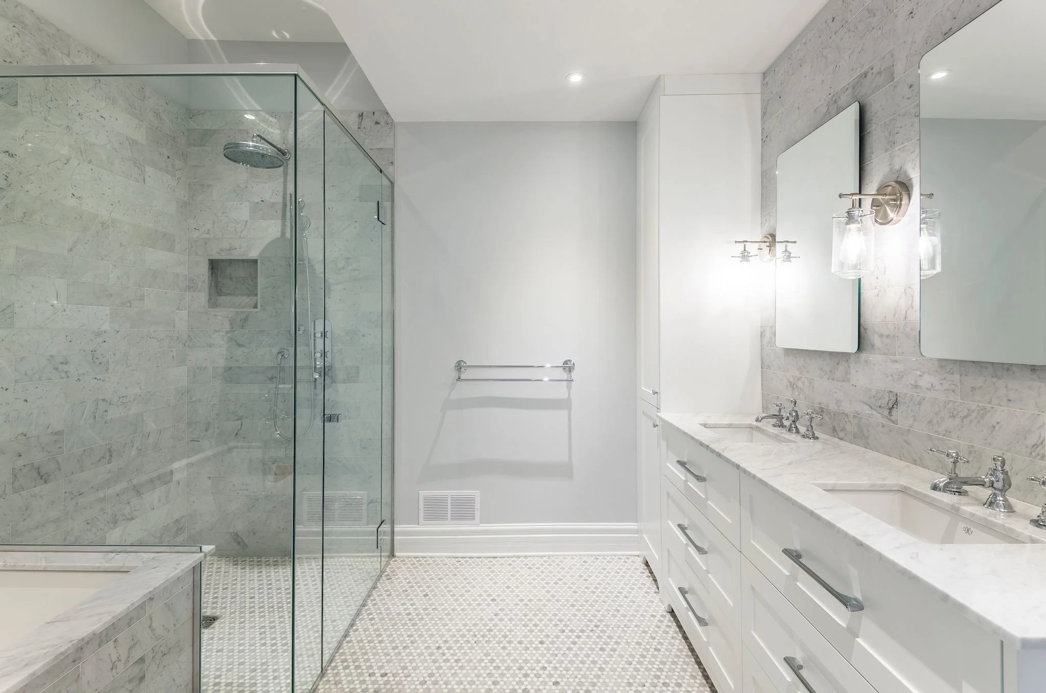 Modern bathroom with a glass-enclosed shower, white marble double vanity with two mirrors, and light fixtures. The floor has small patterned tiles, and the walls are painted light with marble accents.
