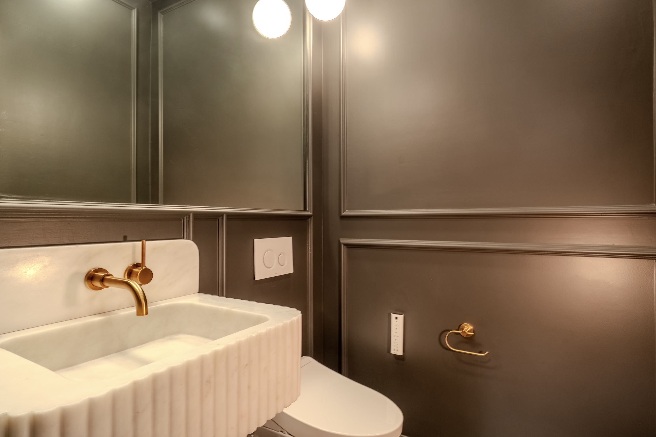 Modern bathroom with dark gray paneled walls, a white marble sink with a gold faucet, and an illuminated mirror above the sink.