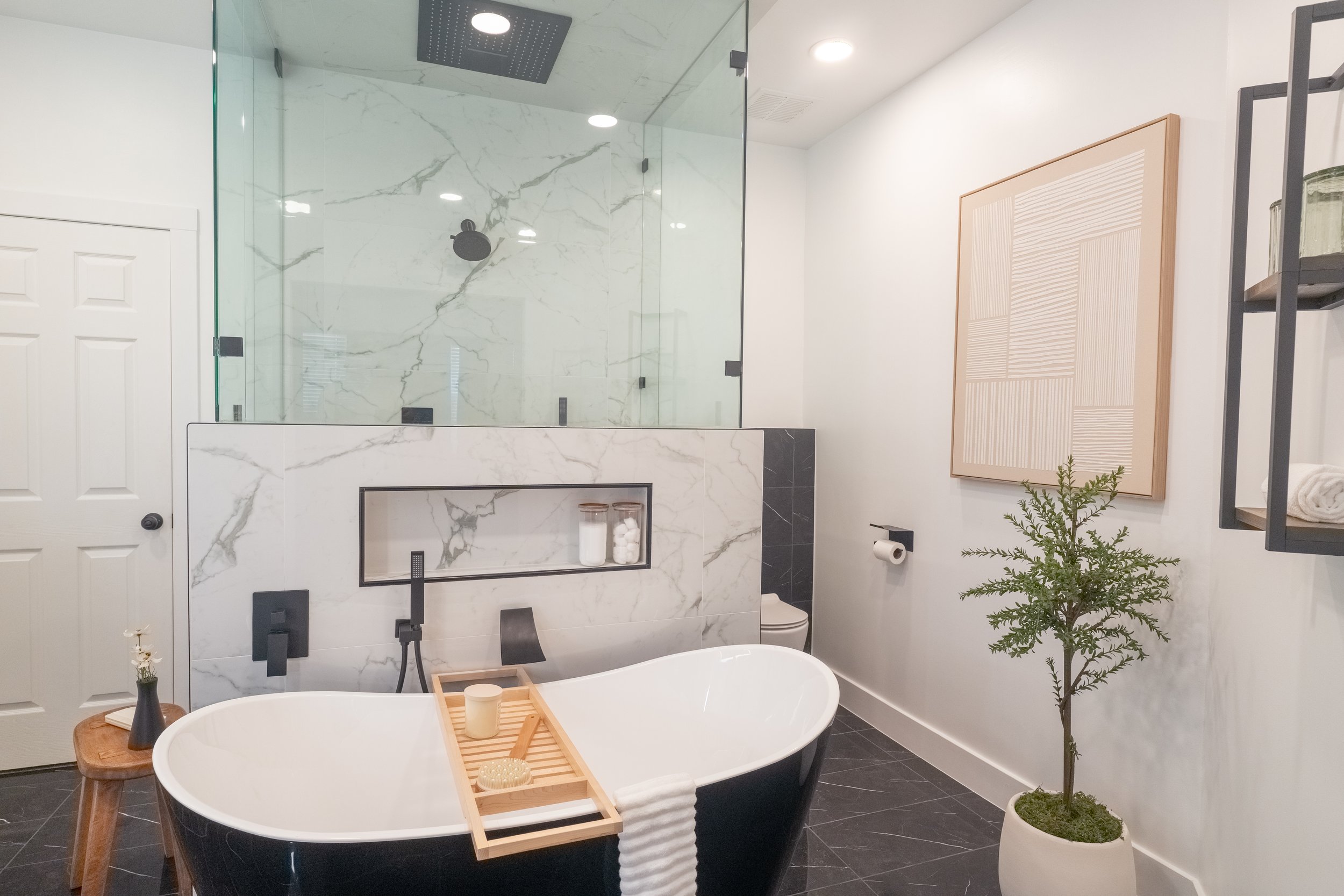 Modern bathroom with black bathtub, wooden tray, white marble walls, glass shower, potted plant, artwork, and minimalist design.