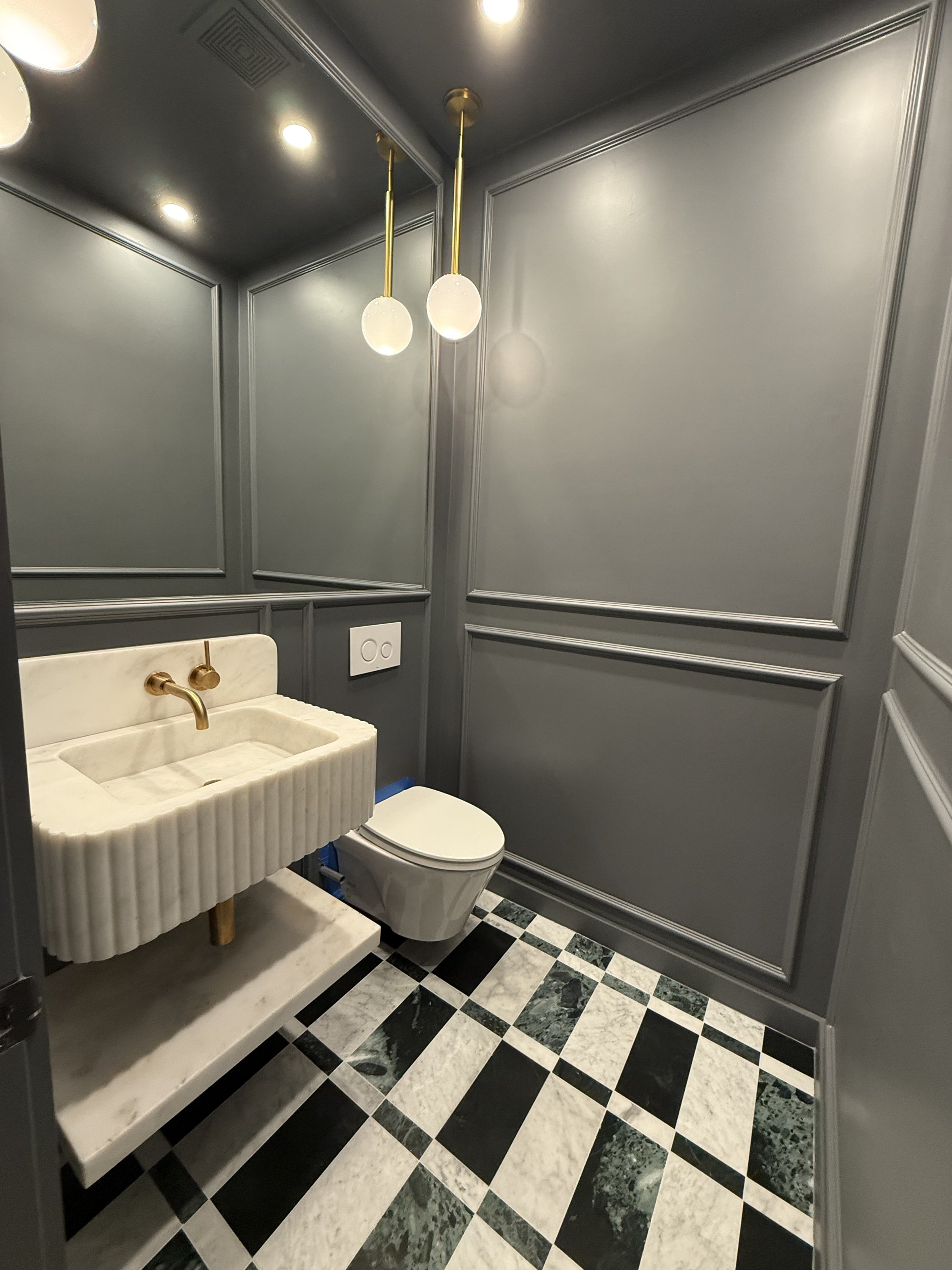 Modern gray-paneled bathroom with a small marble sink with gold fixtures, a wall-mounted toilet, black and white patterned marble tile floor, and two pendant lights hanging from the ceiling.
