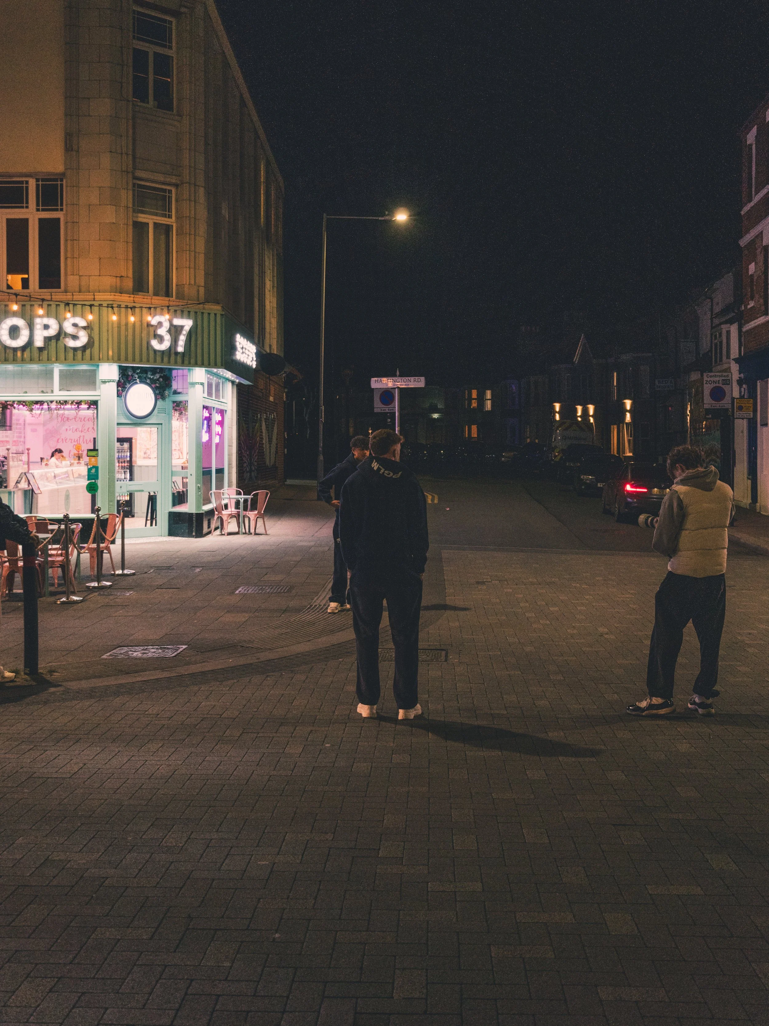 Night scene of a street corner with three people standing on the sidewalk, a brightly lit pink storefront on the left, and parked cars in the background.