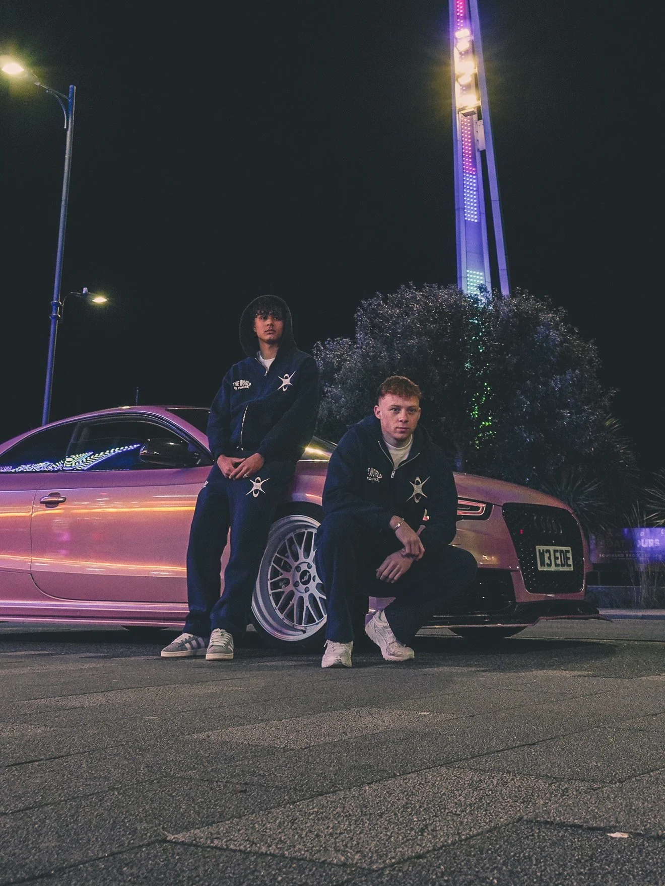 Two young men posing in front of a pink Audi car at night with a brightly lit tower in the background.