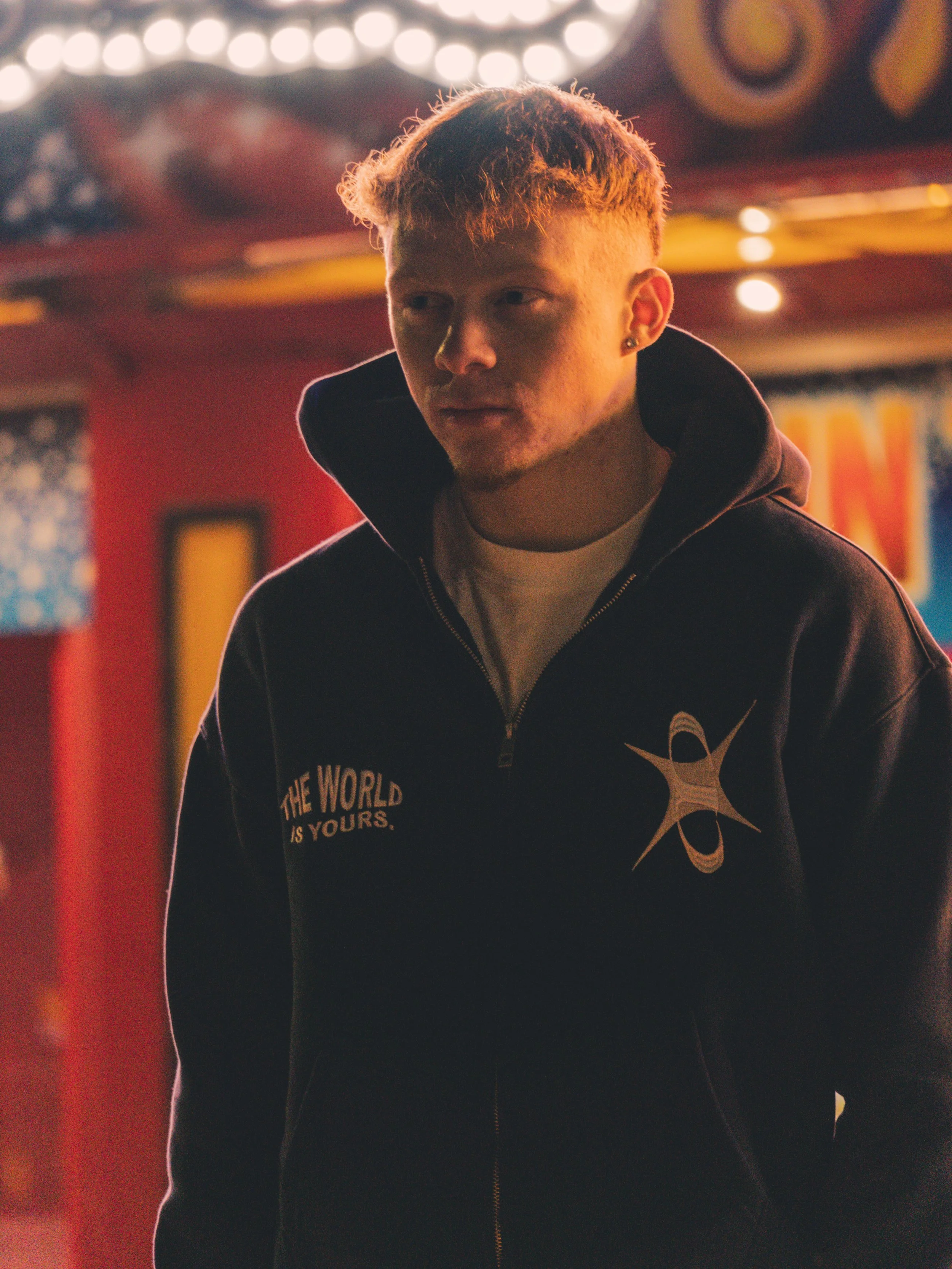 A young man with reddish hair wearing a black zip-up hoodie with a logo and text, standing indoors under colorful lights.