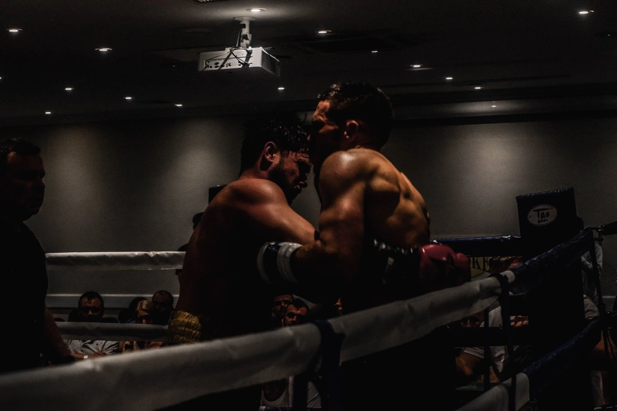 Two shirtless male boxers in a ring, embracing after a match, with an audience and a referee in background.